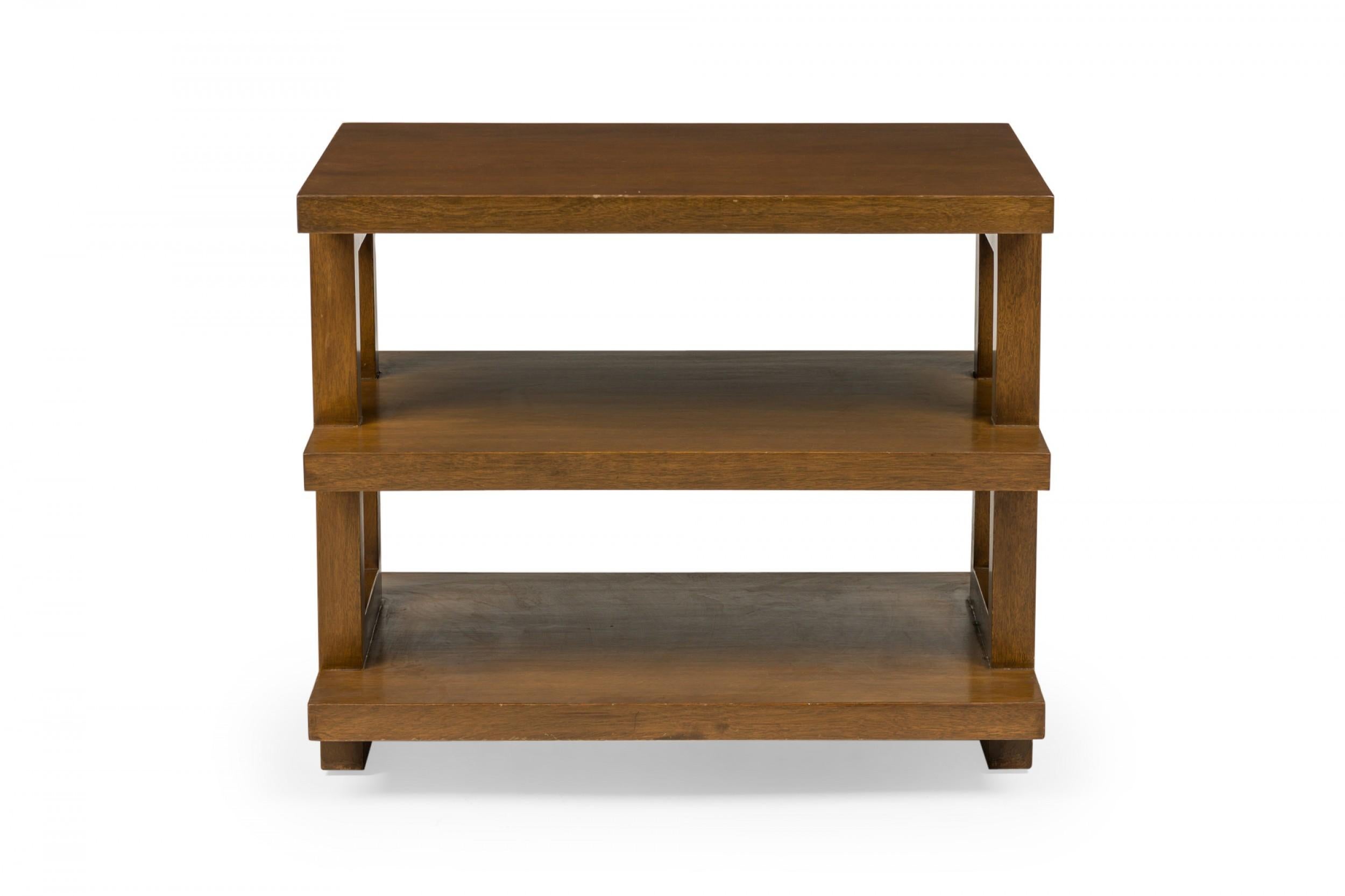 American mid-century three-tier medium brown finished wooden end table with a rectangular top and two shelves below, with rectangular supports on either side, resting on a pedestal base. (Edward Wormley for Dunbar Furniture Company)(Similar tables: