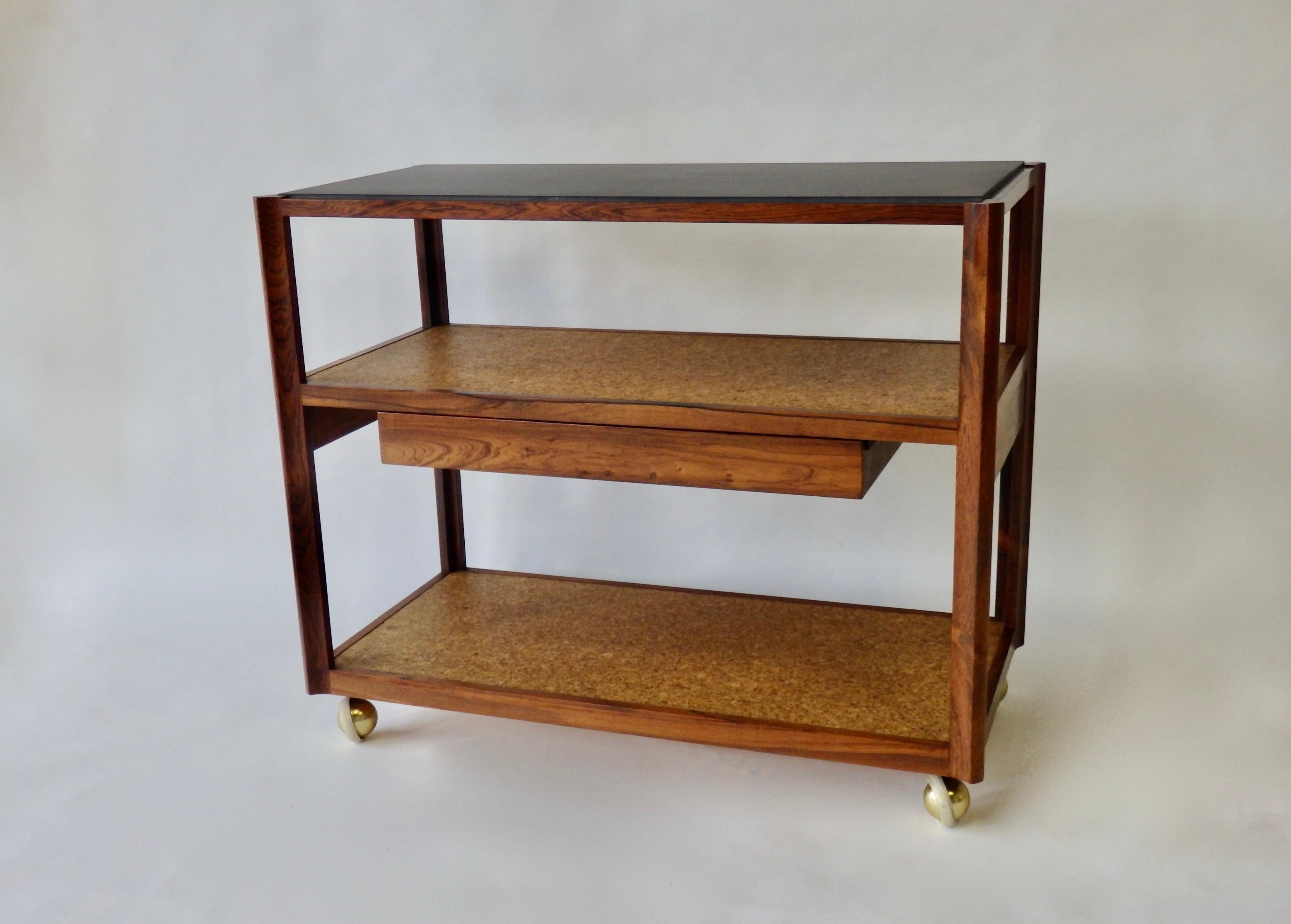 Beautiful Dunbar service or tea cart. Nicely grained rosewood frame supports two cork shelves below slate top. One drawer in mid section shows big D logo. Paper tag remains on underside.