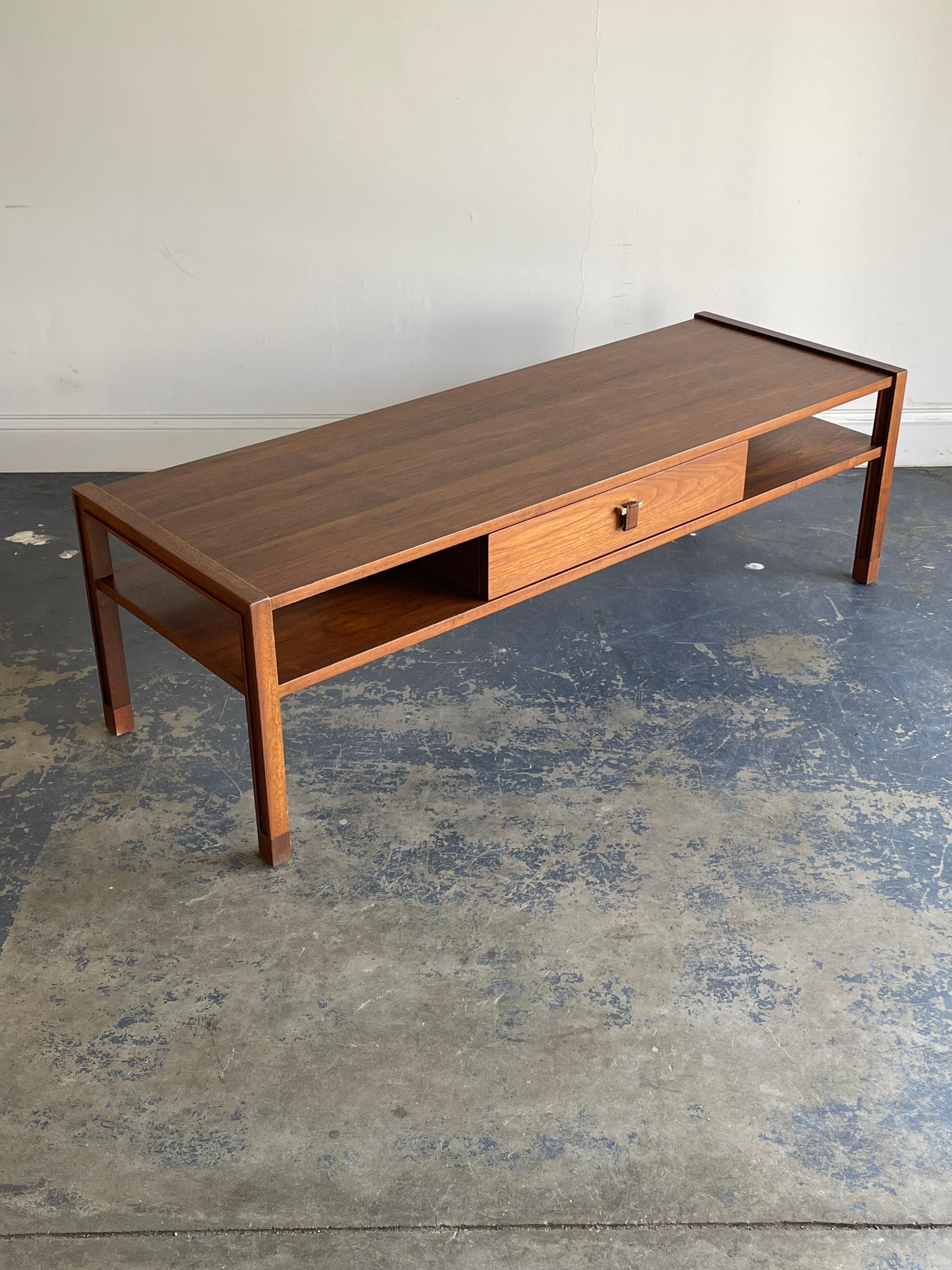 Elegant coffee table designed by Edward Wormley for Dunbar. Table features a rectangular shape with unique leg detail. Below are two open shelves on either side of a drawer accented with brass hardware and a rosewood drawer pull.