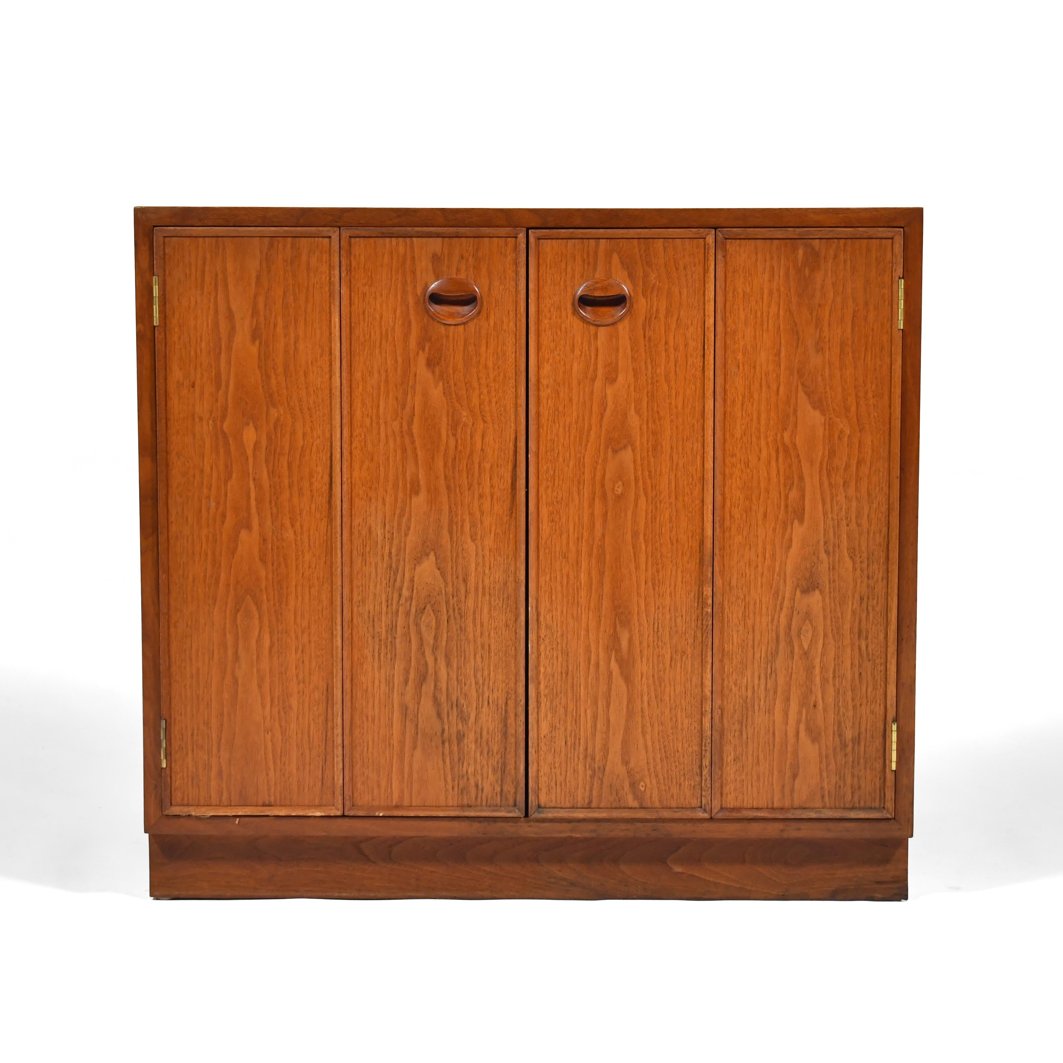 Simple, clean-lined and versatile, this Ed Wormley cabinet by Dunbar features two bi-fold doors concealing one compartment with two shelves. Subtle details include the beautifully figured walnut grain, brass piano hinges in the doors, and the