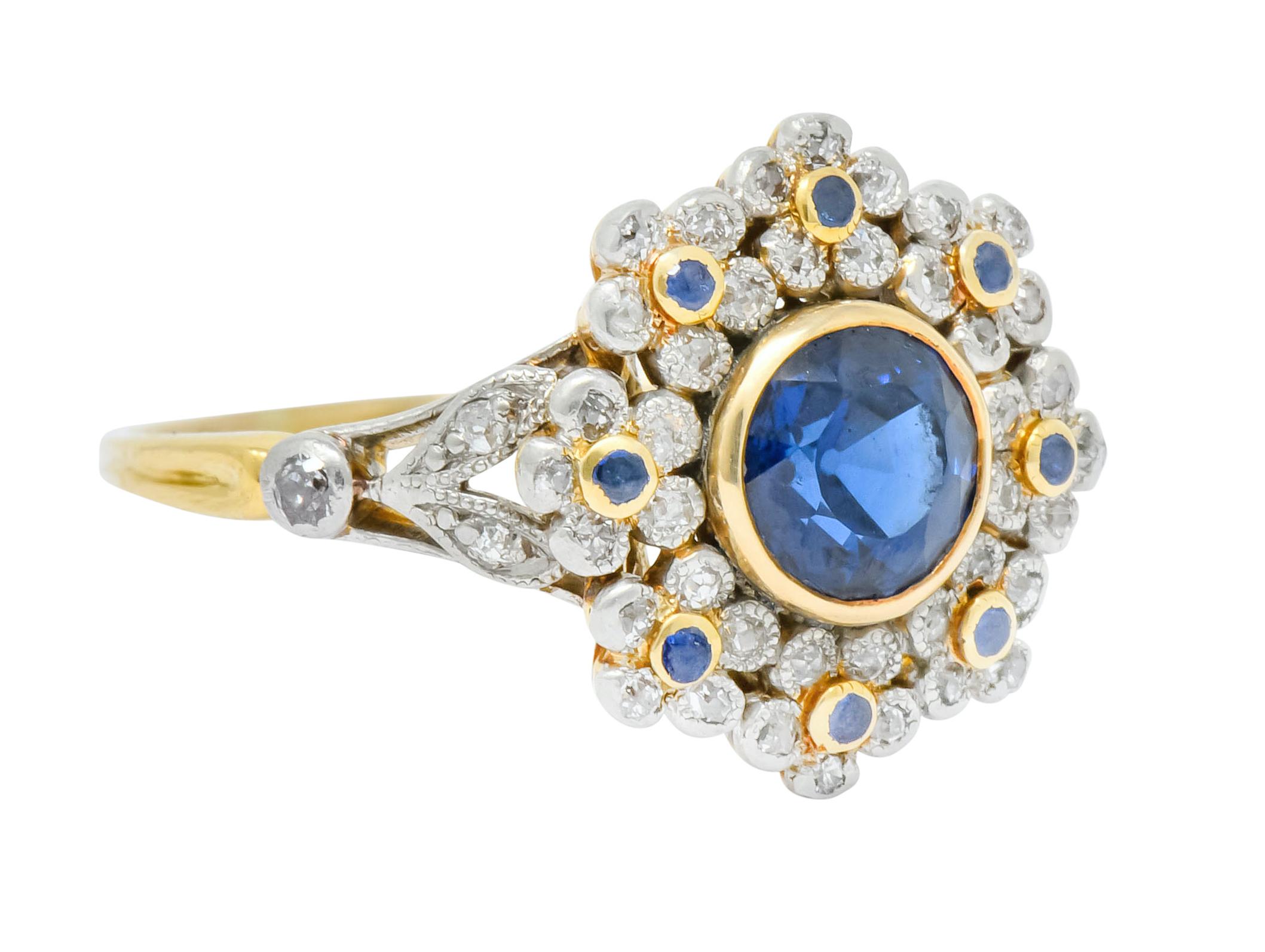 Centering a bezel set round cut sapphire weighing approximately 1.45 carats total, medium-dark royal blue in color

Surrounded by flowers in a cluster style, flanked by platinum foliate split shoulders

Accented throughout by round cut sapphires and