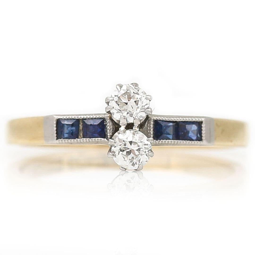 A very pretty early 20th century 18ct gold and platinum, old cut diamond two stone set with sapphire shoulders, dating from circa 1915. The old mine cut stones weighting approx 0.15ct set atop the ring are nestled along side stone another. To the
