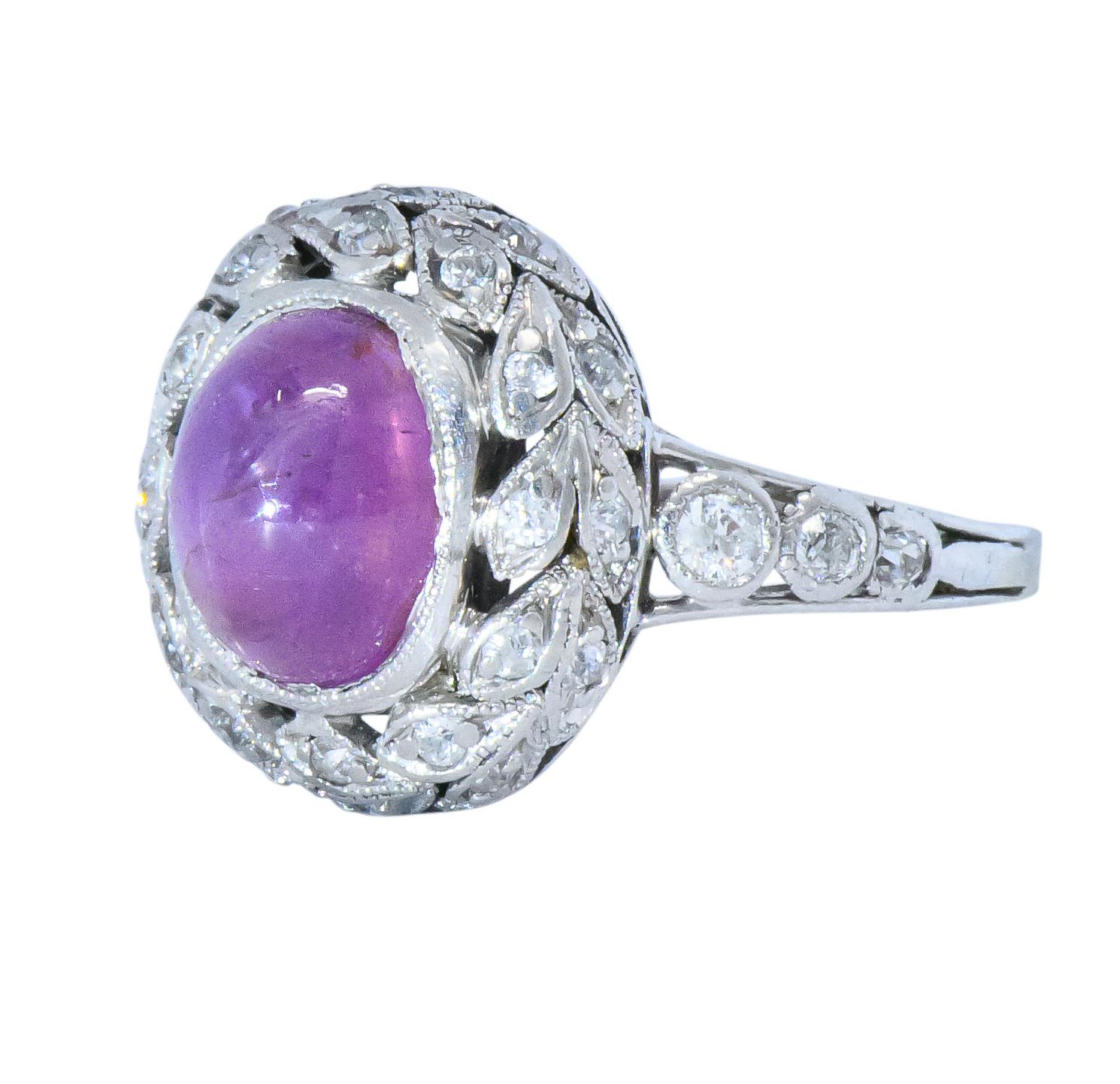 Centering an oval cabochon star pink sapphire weighing approximately 3.35 carats

Translucent and bright purplish-pink with a natural six rayed star 

Bezel set in a pierced mounting with a foliate surround accented by miligrain

Set throughout by