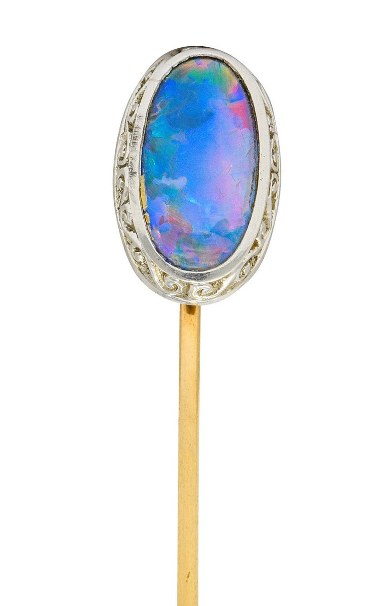 Centering an oval cabochon of black opal, gray in body color with strong blue/red play-of-color and moderate green flash

Bezel set in a platinum surround decorated with a scrolled motif

Completed by a 14 karat gold pin stem

Circa: 1900

Top