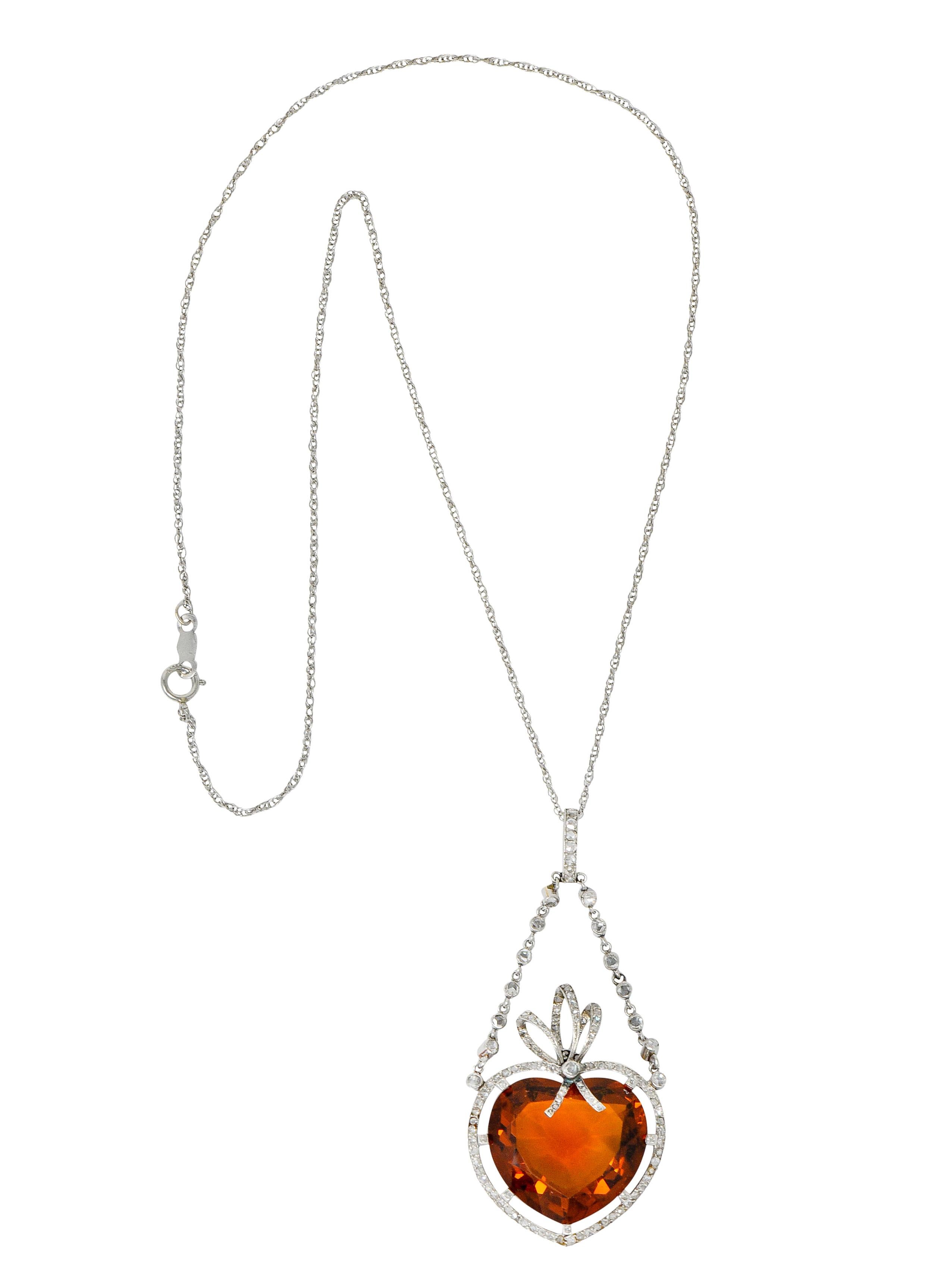 Centering a mixed heart cut citrine weighing approximately 12.00 carats, transparent and medium-dark with slightly brownish-orange color

Surrounded by a diamond halo that terminates as a ribboned bow at top of heart, suspending from two bezel link