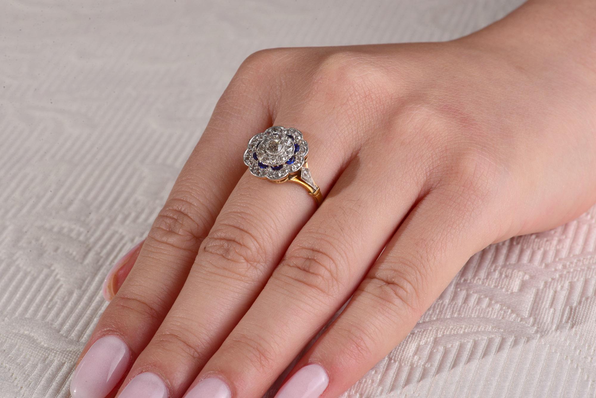 Edwardian cluster ring with a centre old cut diamond of 1 x 0.02 cts surrounded by 11 1.5mm approximately old single cut diamonds of 1.5 to 1.7mm framed by half moon grain set Synthetic Sapphires. The outer scalloped circle of the ring is set with