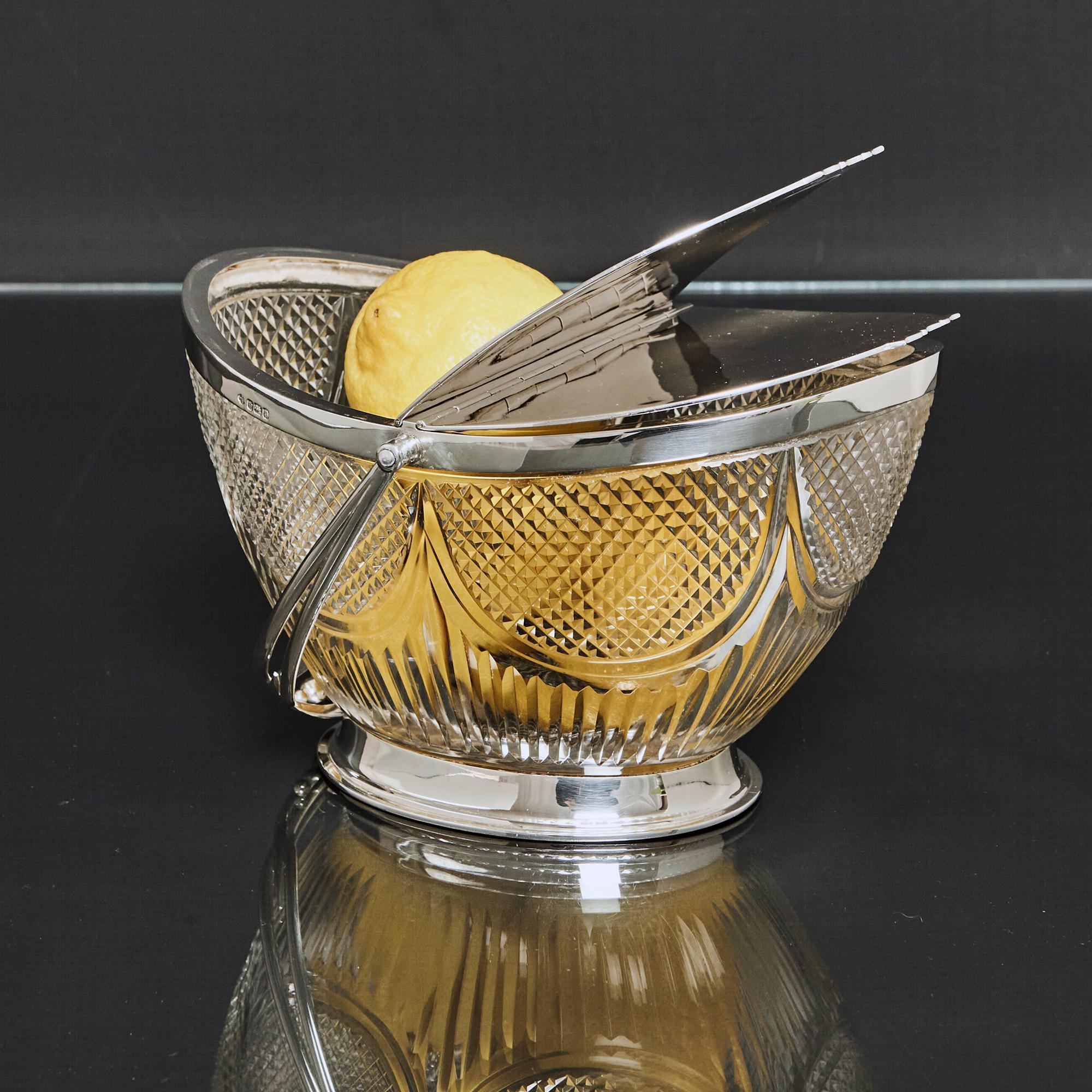 Edwardian, antique cut glass box of oval, boat-shaped form - a fruit basket or biscuit box. The cut glass body is decorated with elliptical panels of a crisply cut diamond pattern, and vertical fluting. The body is fitted with silver mounts, a swing