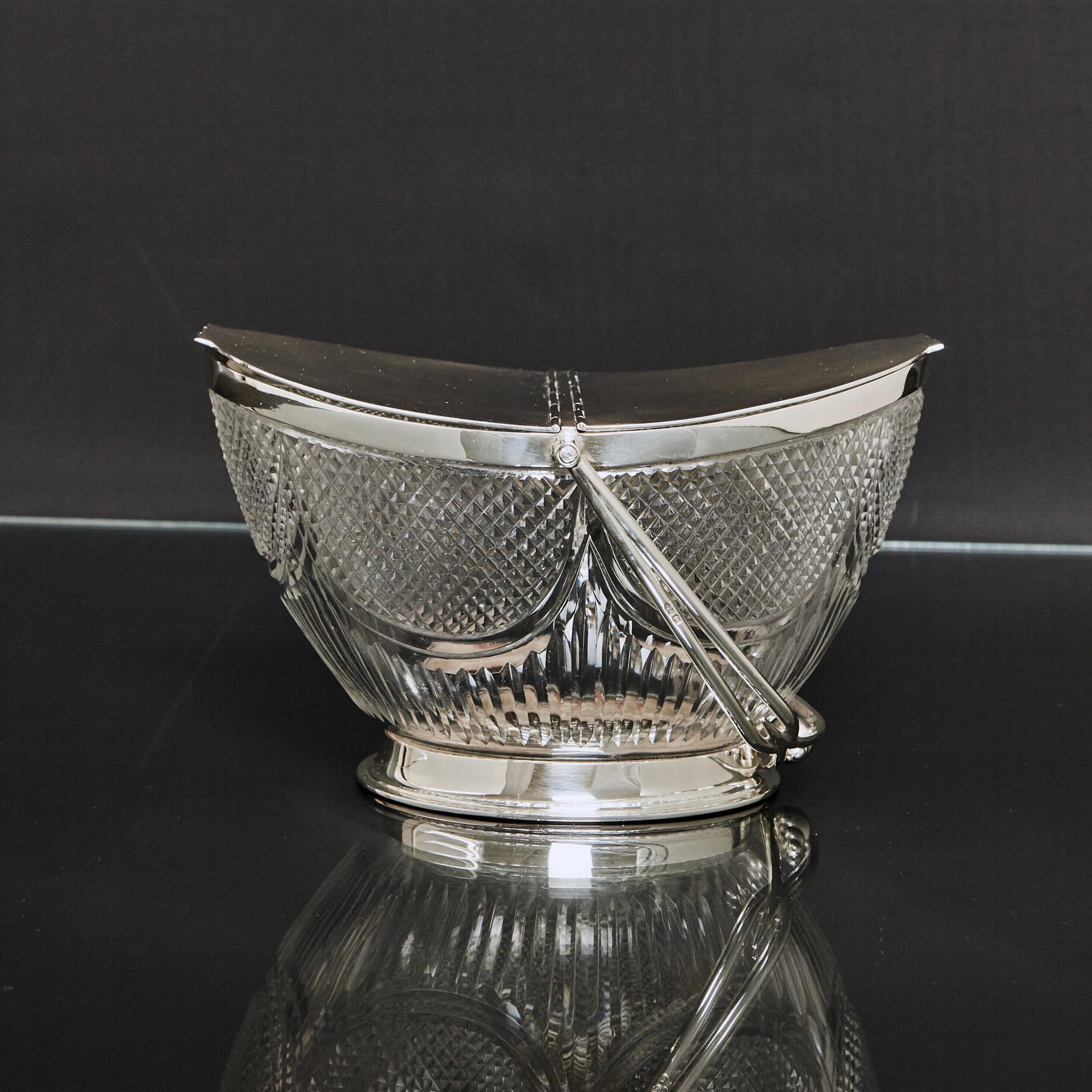 Edwardian, antique cut glass box of oval, boat-shaped form - a fruit basket or biscuit box. The cut glass body is decorated with elliptical panels of a crisply cut diamond pattern, and vertical fluting. The body is fitted with silver mounts, a swing