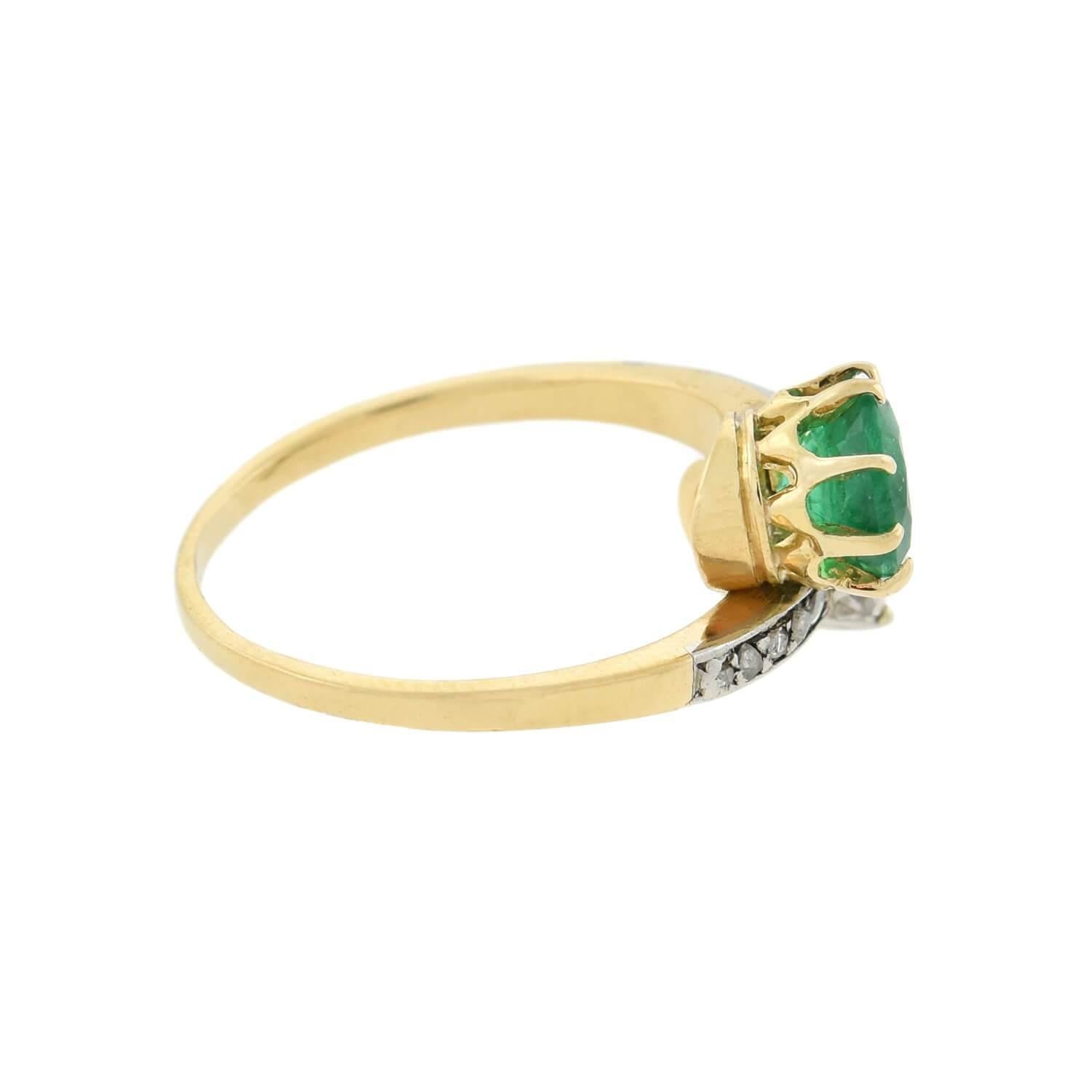 A unique and exquisite diamond bypass ring from the Edwardian (ca1910) era! Crafted in 18kt yellow gold topped with platinum, this piece features an Old European Cut diamond and a vibrant green Round emerald nestled together at the center, forming a