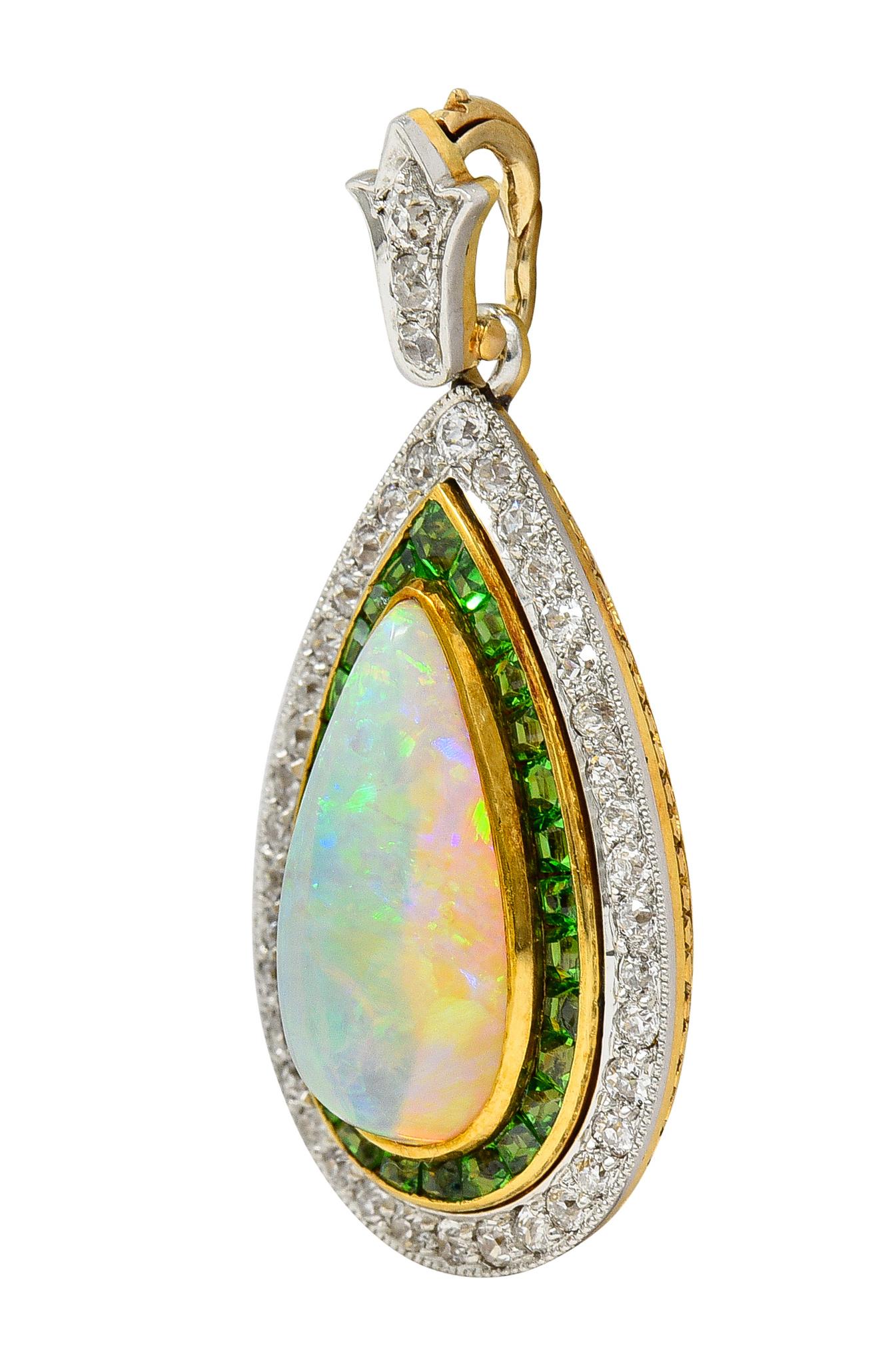 Comprised of a lily motif surmount suspending a teardrop-shaped pendant centering an opal cabochon
Pear shaped and measuring 7.0 x 15.0 mm - white in body color with spectral play-of-color
Set in a gold bezel with a double halo surround of demantoid