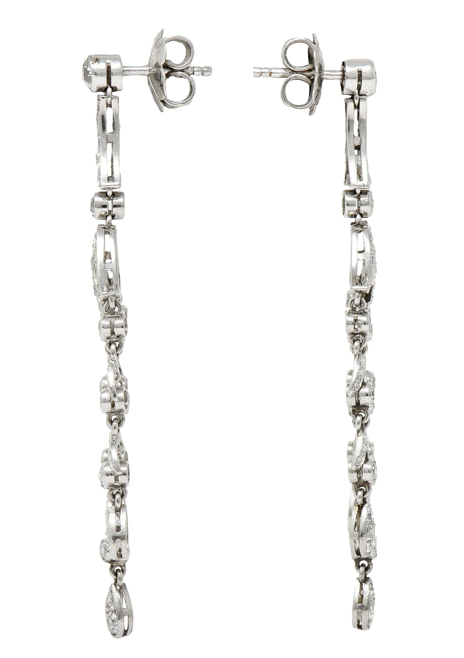 Chandelier style earrings have diamond stud surmounts. Suspending a bar link that extends into a flourished foliate motif. With three articulated tendrils of bezel set diamond links alternating with diamond accented foliate. Total diamond weight is