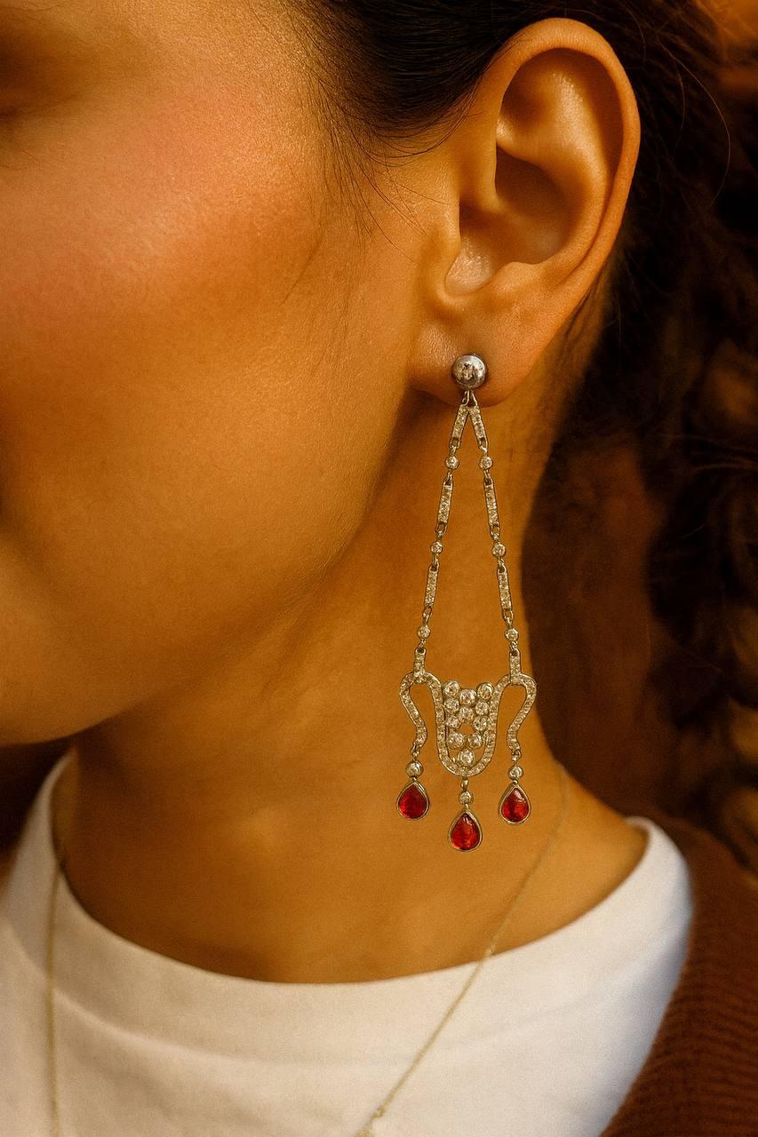 Magnifique paire de boucles d'oreilles chandelier de style édouardien en or blanc 18 carats, présentant une élégante composition de diamants naturels et de rubis éclatants.
Le motif ajouré complexe brille avec environ 5,70 carats de diamants,