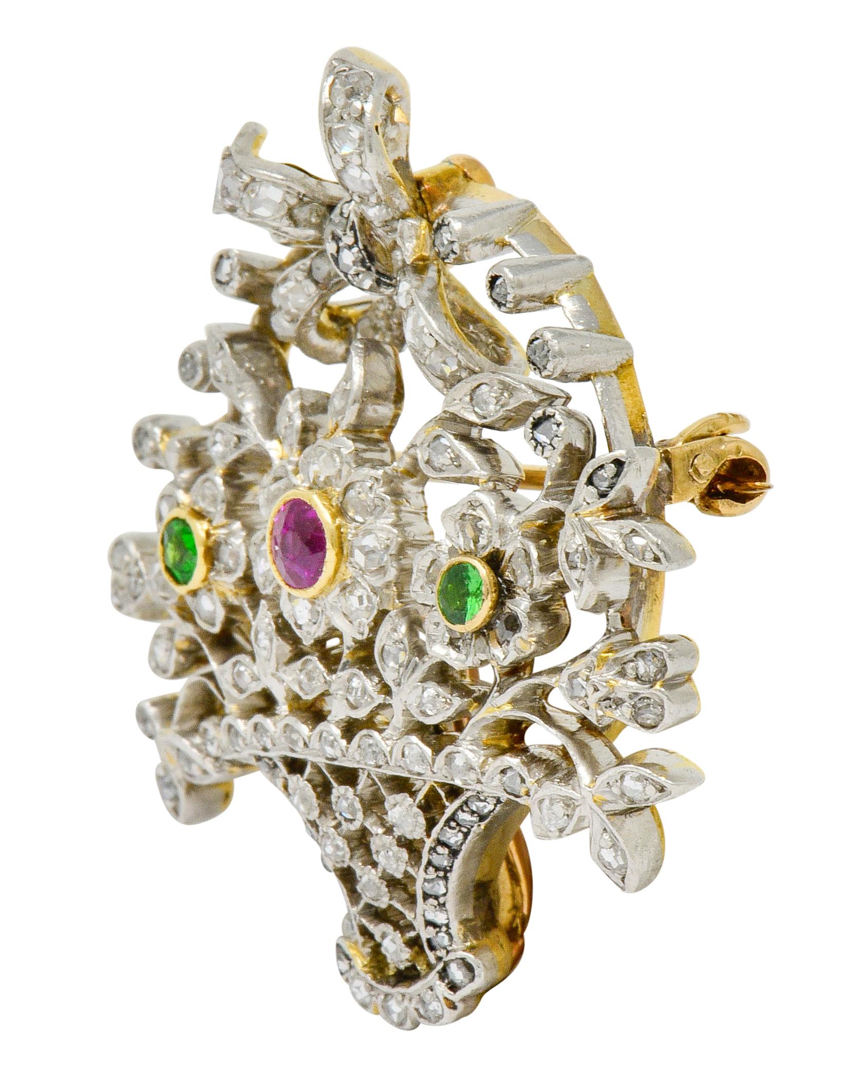 Brooch is designed as a trellised basket overflowing with burgeoning florals and foliate

Centering a round cut ruby, bezel set in yellow gold, weighing approximately 0.18 carat; bright purplish-red in color

Flanked by two round cut demantoid