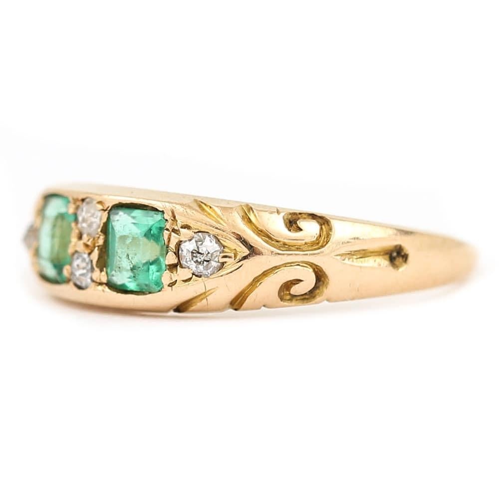 A pretty Edwardian 18 karat gold emerald and diamond boat shaped cluster ring. With two cushion cut emeralds each estimated at 0.15ct each surrounded by four old cut diamonds in a carved/scroll setting. The hallmark is worn but it looks as if it was