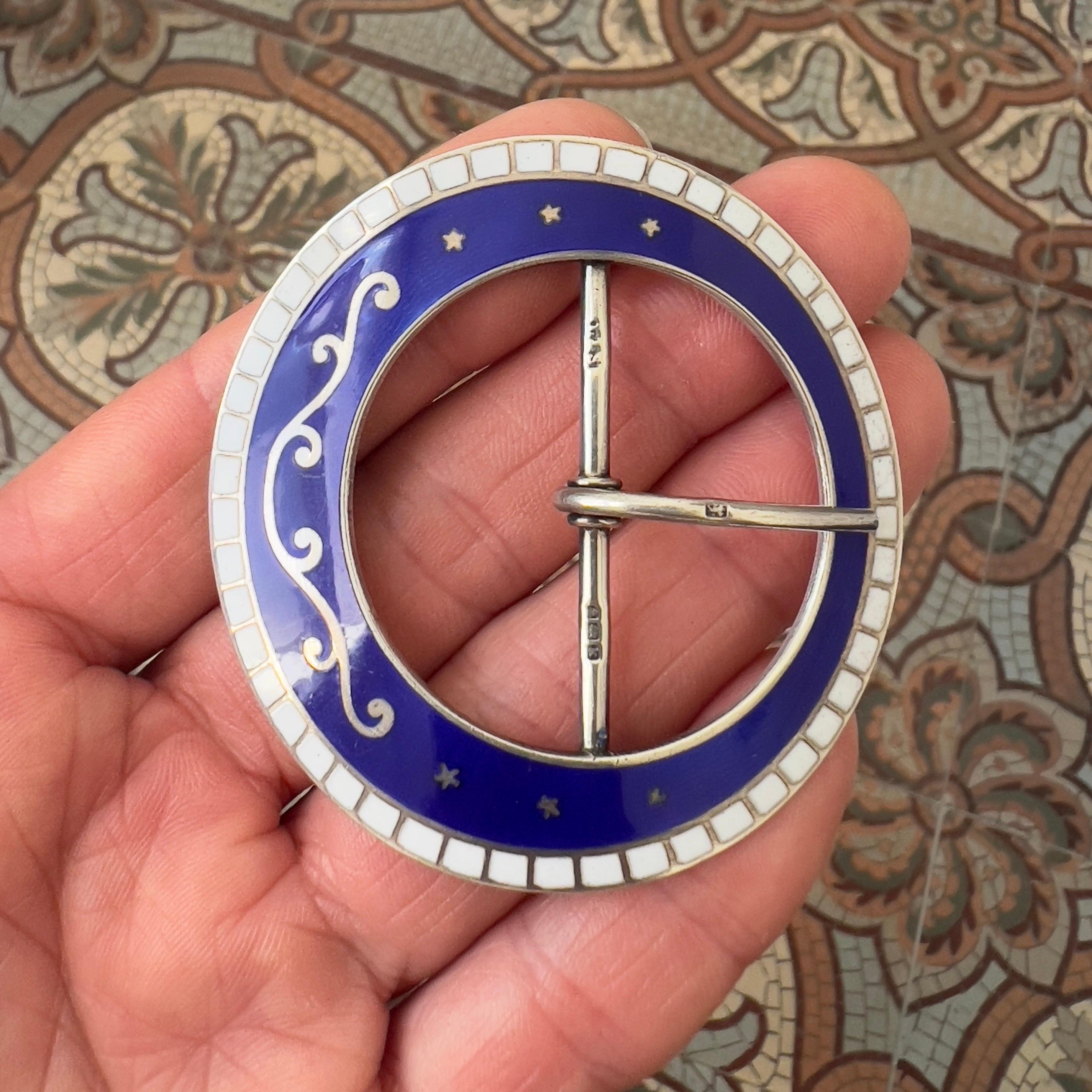 A captivating example of Edwardian craftsmanship, this circular belt buckle is rendered in sterling silver and adorned with deep royal blue enamel, accented by elegant silver scrollwork and a delicate border of white enamel tiles. The celestial