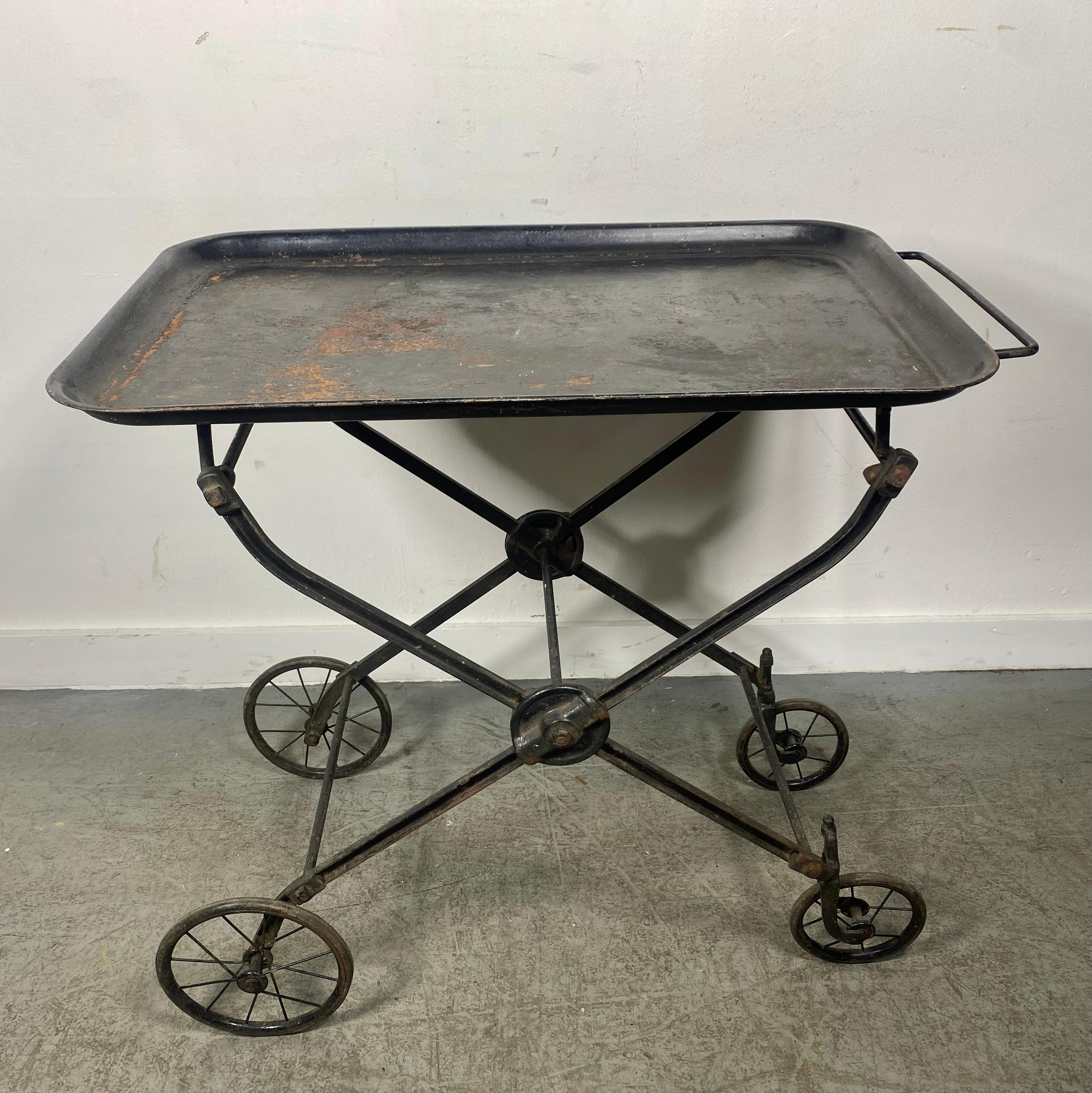 Handled rolling and folding iron hotel beverage trolley, serving cart, dry bar. Tea. Coffee. Plants. Display. Featuring a rectangular larger lipped tray, sturdy Iron crossbar and castings with spoked wheel detail. Uncommon. Industrial. Fine design.