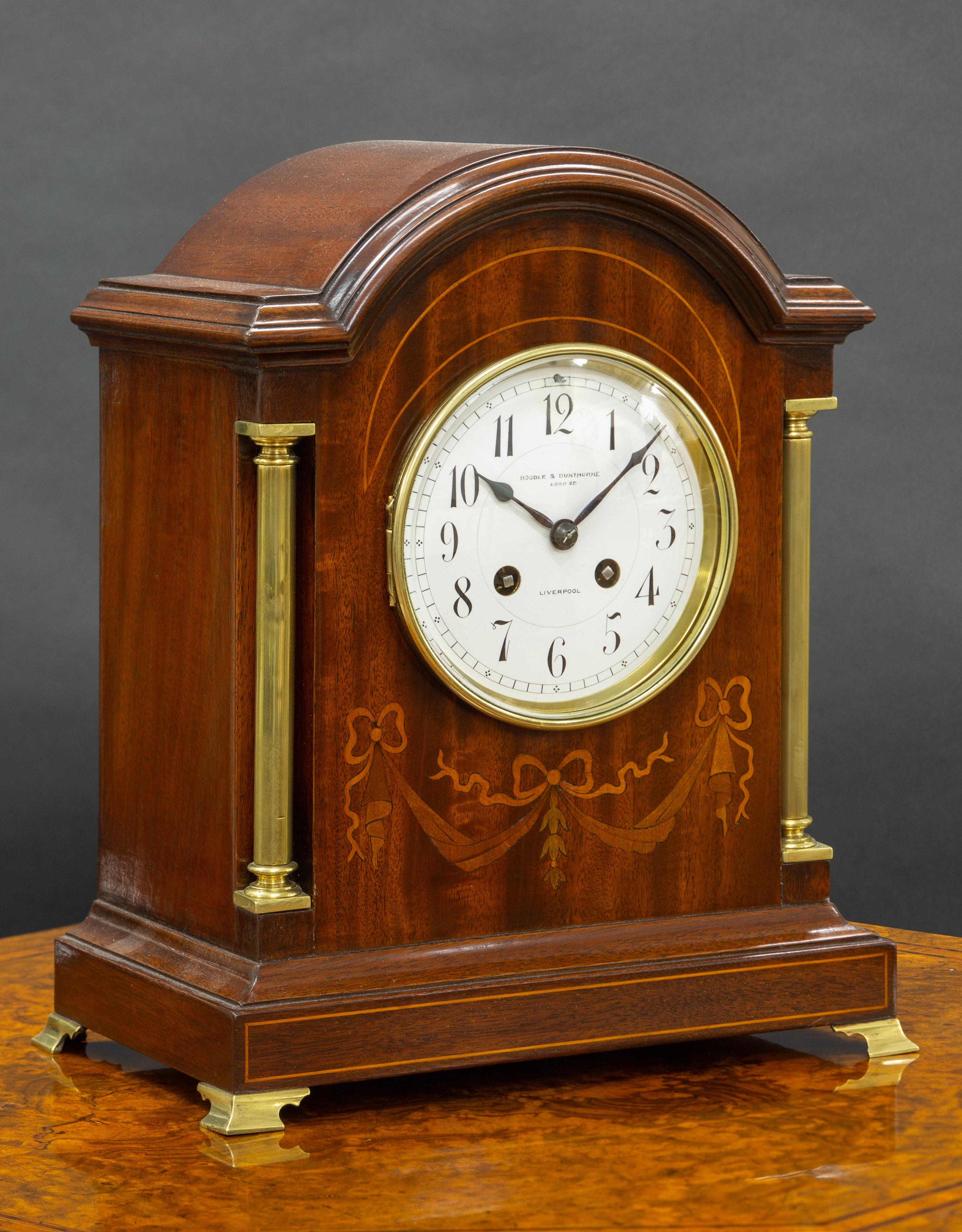 Edwardian Mantel clock in a mahogany case with brass pillars and satinwood floral and line inlaid decoration, standing on brass outswept bracket feet. 

Enamel dial with Arabic numerals, original ‘blued’ steel hands signed ‘Boodle and Dunthorne.