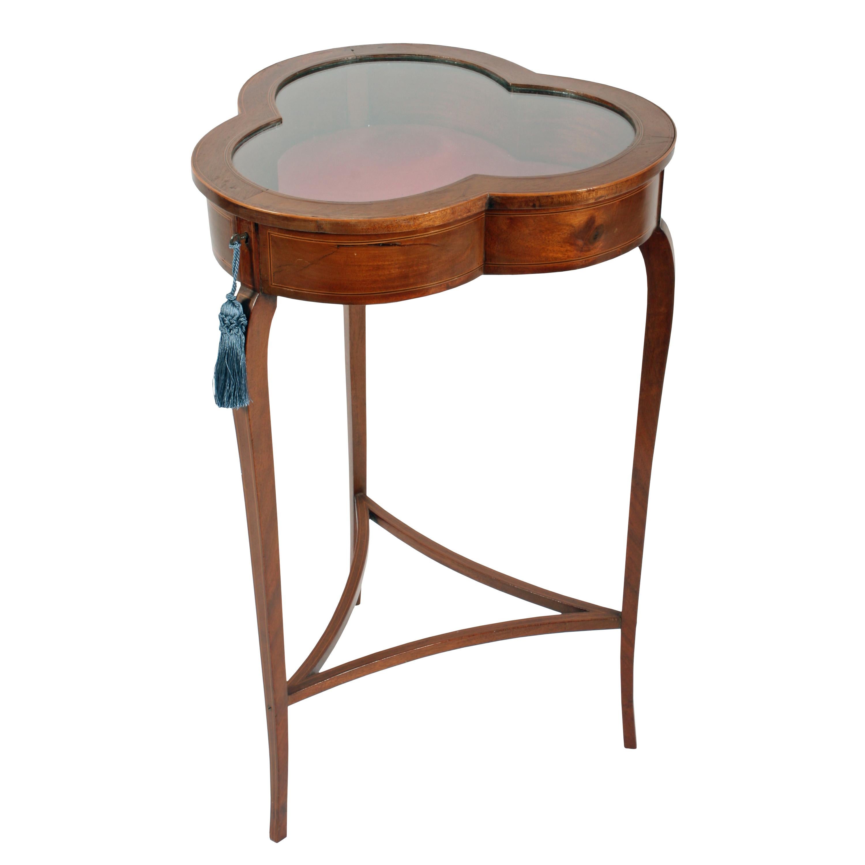 An early 20th century Edwardian inlaid mahogany clover leaf shaped bijouterie table or vitrine.

The shaped top has a glazed hinged lid with a lock and key and a dark pink coloured material interior.

The table stands on three square sided