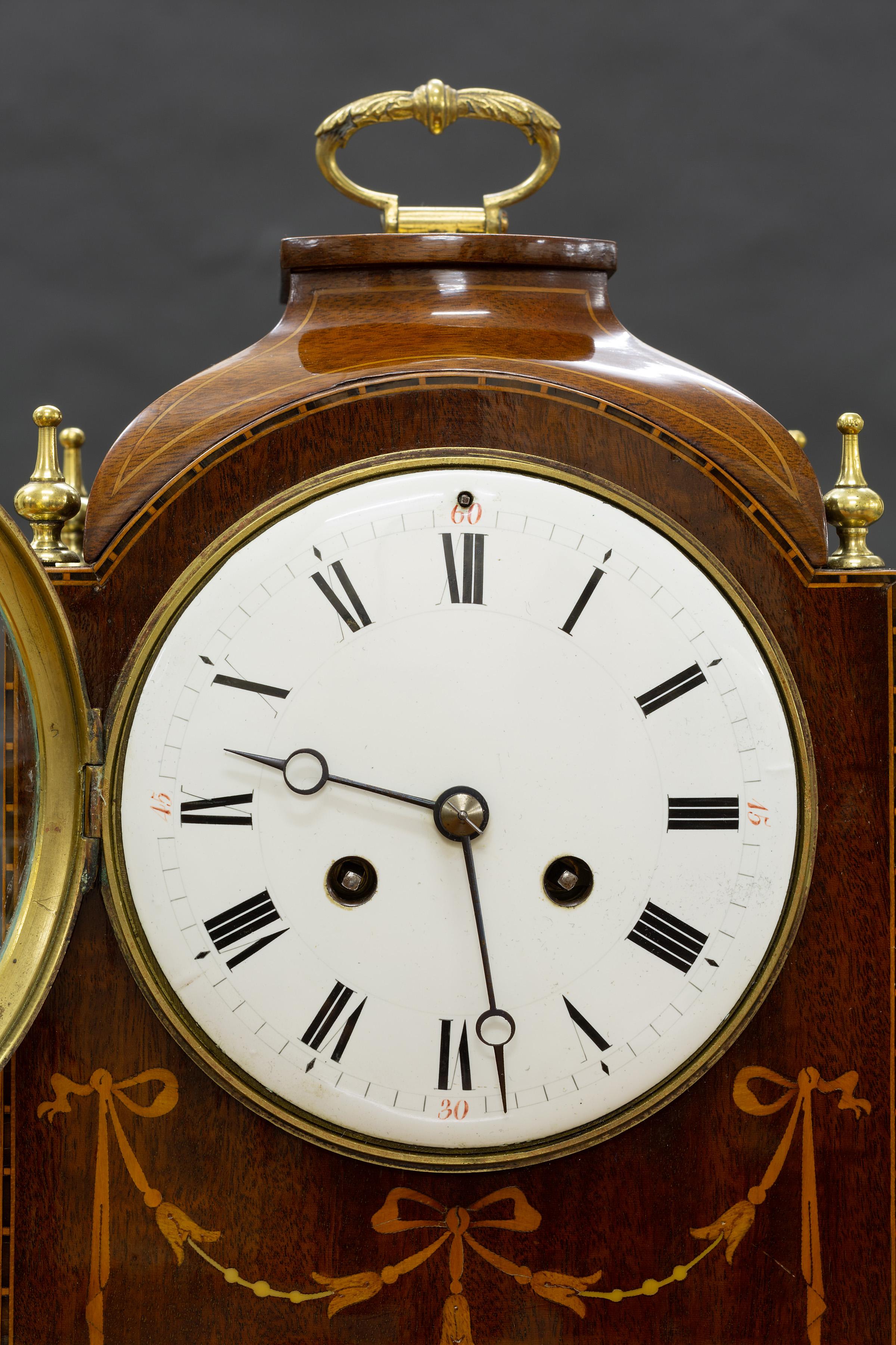 Edwardian mahogany mantel clock standing on brass bracket feet in an arch top case surmounted by four finials with satinwood swag decoration.

Enamel dial with Roman numerals and original ‘blued’ steel hands.

Eight day French movement striking