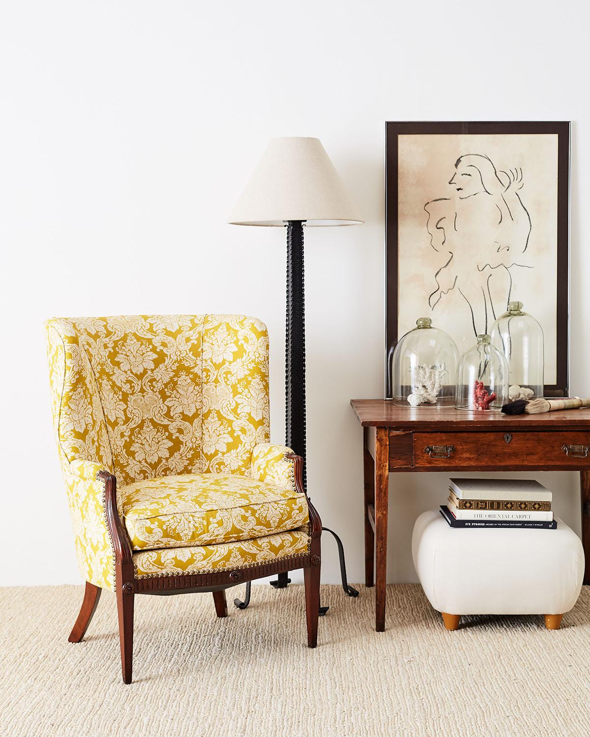 Colorful Edwardian mahogany wing chair featuring a classic yellow print fabric, probably Fortuny. The mahogany frame has molded arms and the upholstery around the front is accented with brass tack nail heads. The serpentine seat is decorated with