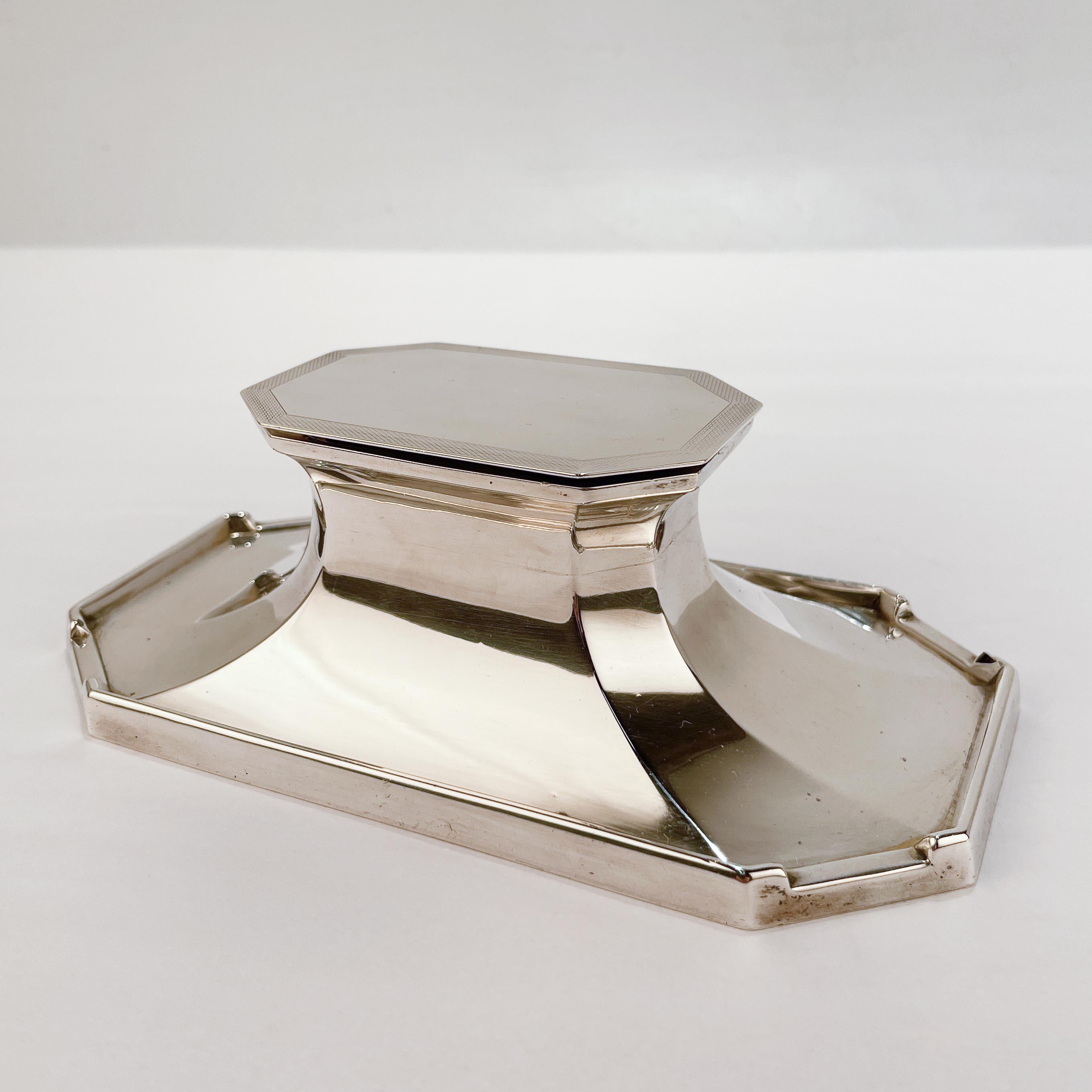 A fine English sterling silver inkwell.

In the Edwardian style. 

With notched corners to the base for pens, a sloped pedestal center that supports two gilt inkwell chambers and a hinged lid with engine-turned decoration.

The inkwell is cement