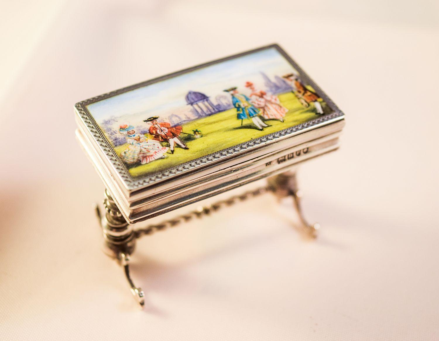 ENAMEL MINIATURE SILVER TABLE £425      £1200

Edwardian miniature silver and enamel table box. 
Modelled as an occasional
table on turned supports joined bay a stretcher.
The enamel top with 18th century figures in a landscape.
London import marks