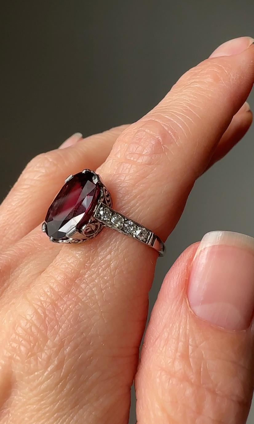 Exquisite in every way, this regal Edwardian ring centers on a glowing 7.8 carat plum-colored natural spinel. The mounting is like a tiny crown with diamond lined shoulders, finished with delicate hand engraving and elegant openwork. Currently a