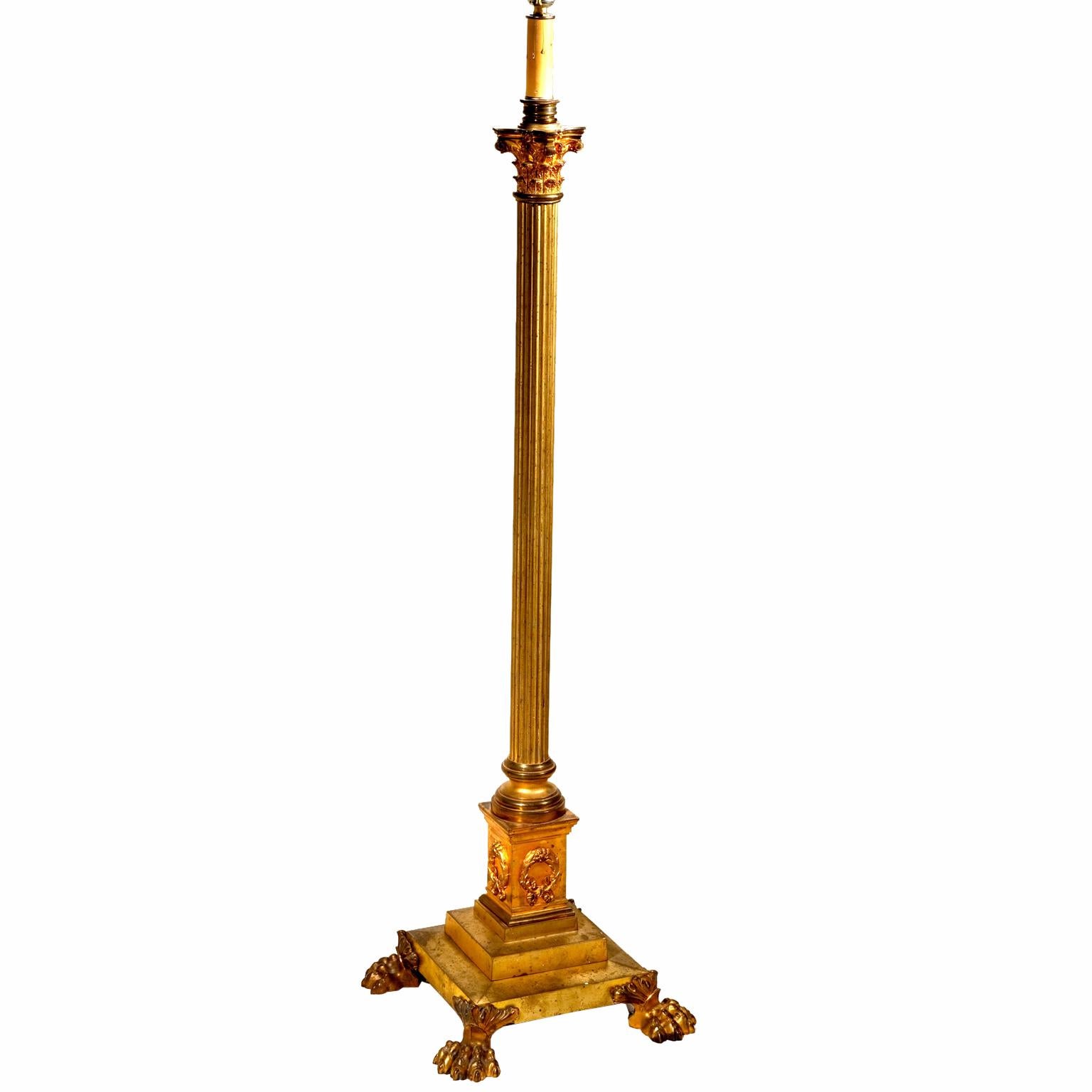 A neoclassical gilded brass floor lamp with a stepped base on four lion’s paw feet; the central fluted column resting on a rectangular plinth decorated with wreaths, the top of the column terminates in a Corinthian capital; the lamp comes complete