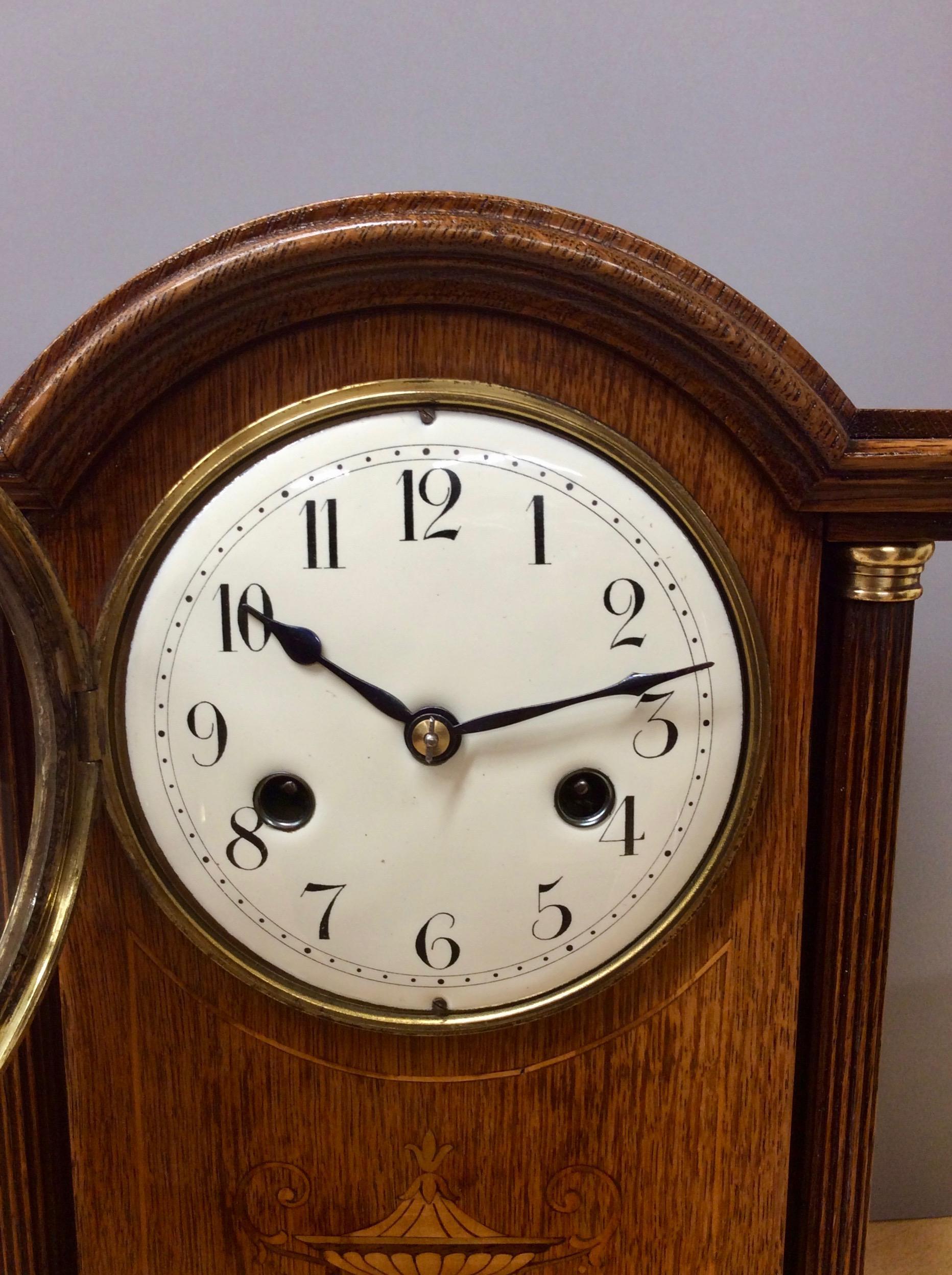 Edwardian oak cased mantel clock standing on a stepped base with bun feet. Turned pillars with brass capitals either side of a beautifully decorated satinwood inlay depicting an urn.

Enamel dial with Arabic numerals and original steel