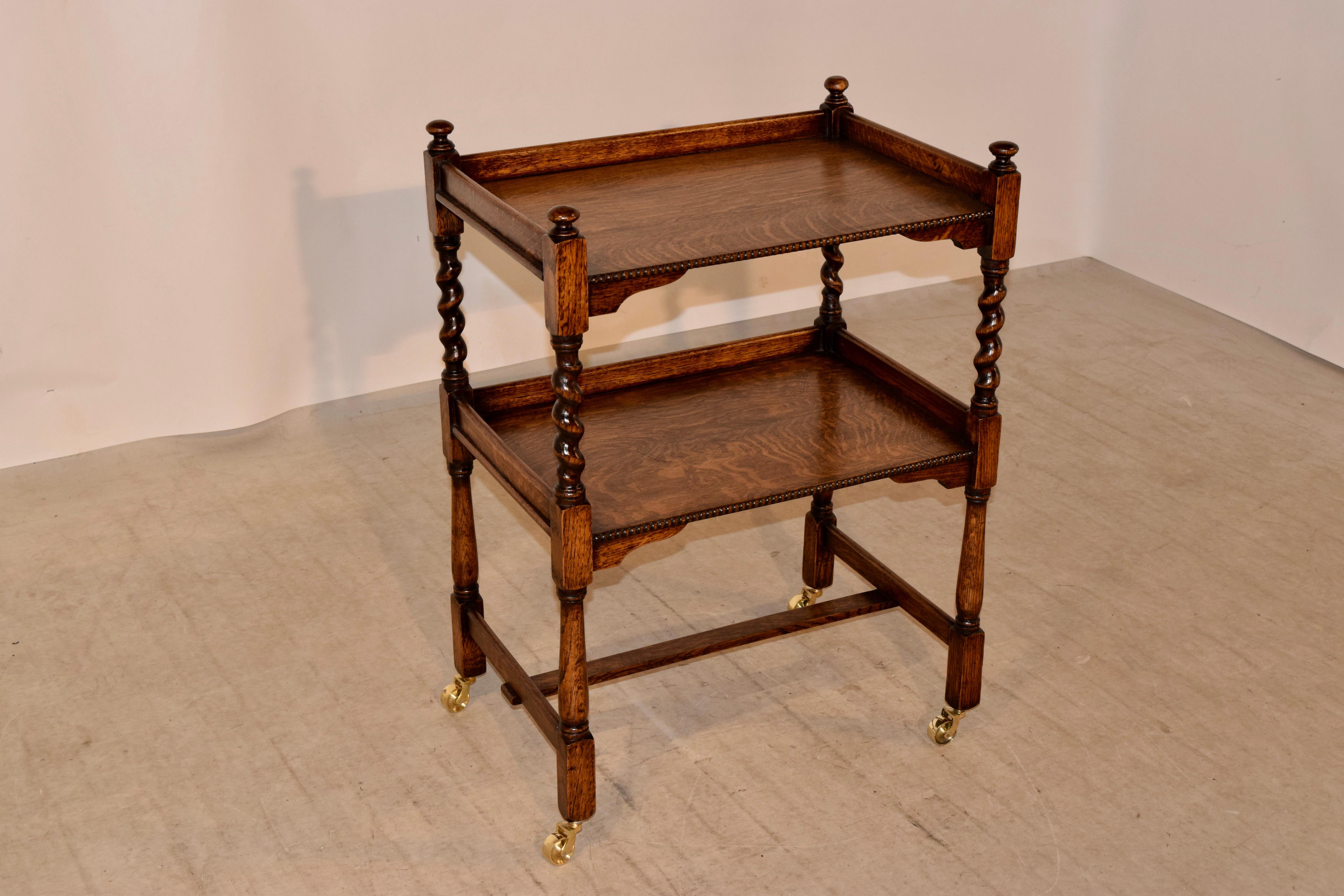 Edwardian drinks cart from England made from oak with two shelves which have beaded edges across the front, separated by hand-turned barley twist shelf supports following down to hand-turned legs joined by simple stretchers. Casters are hand cast