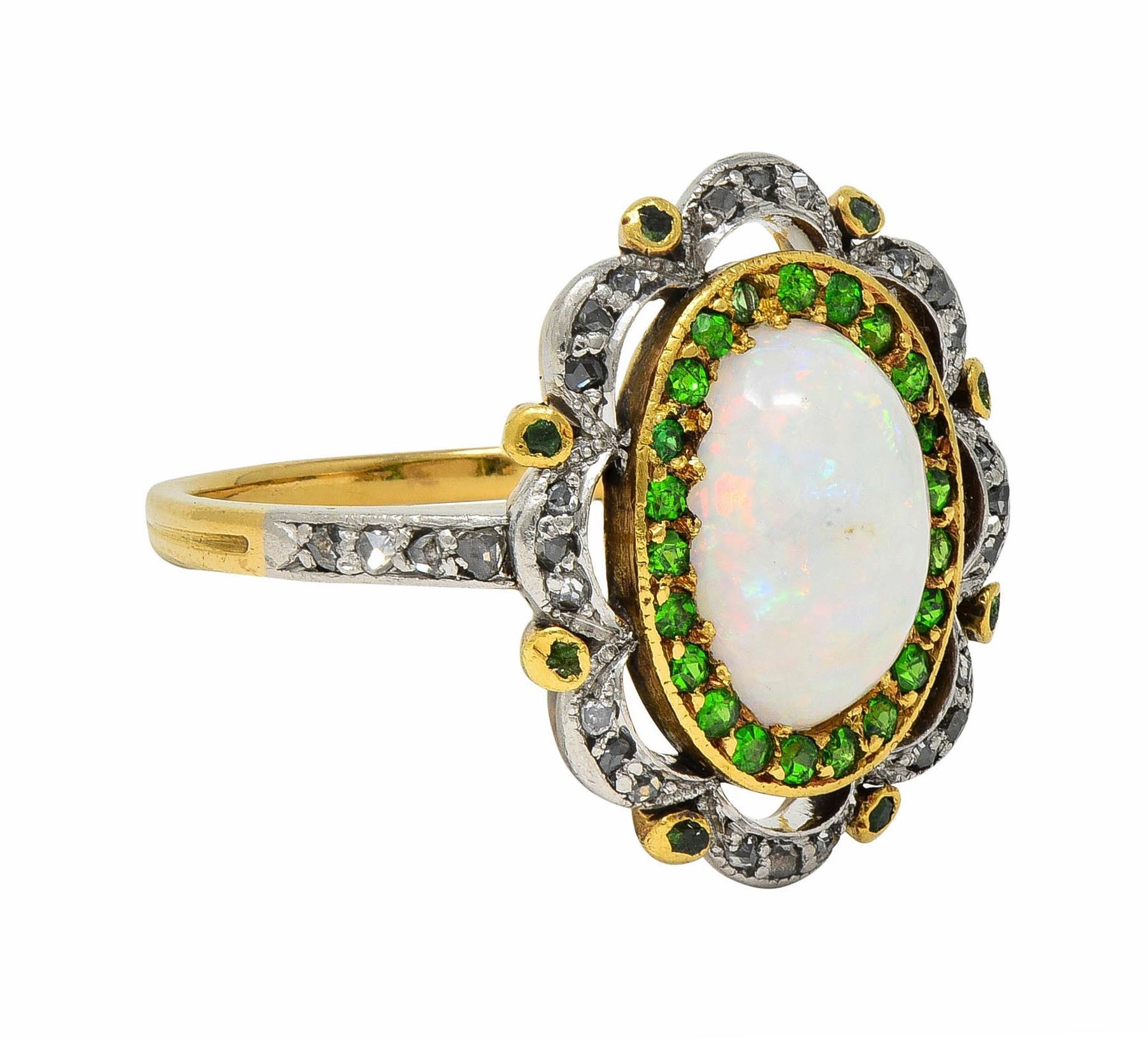 Centering an oval-shaped opal cabochon - white in body color with spectral play-of-color
Measuring 6.5 x 9.5 mm - bead set in yellow gold with halo surround of demantoid garnets 
Round cut and bead set with additional granter bezel set