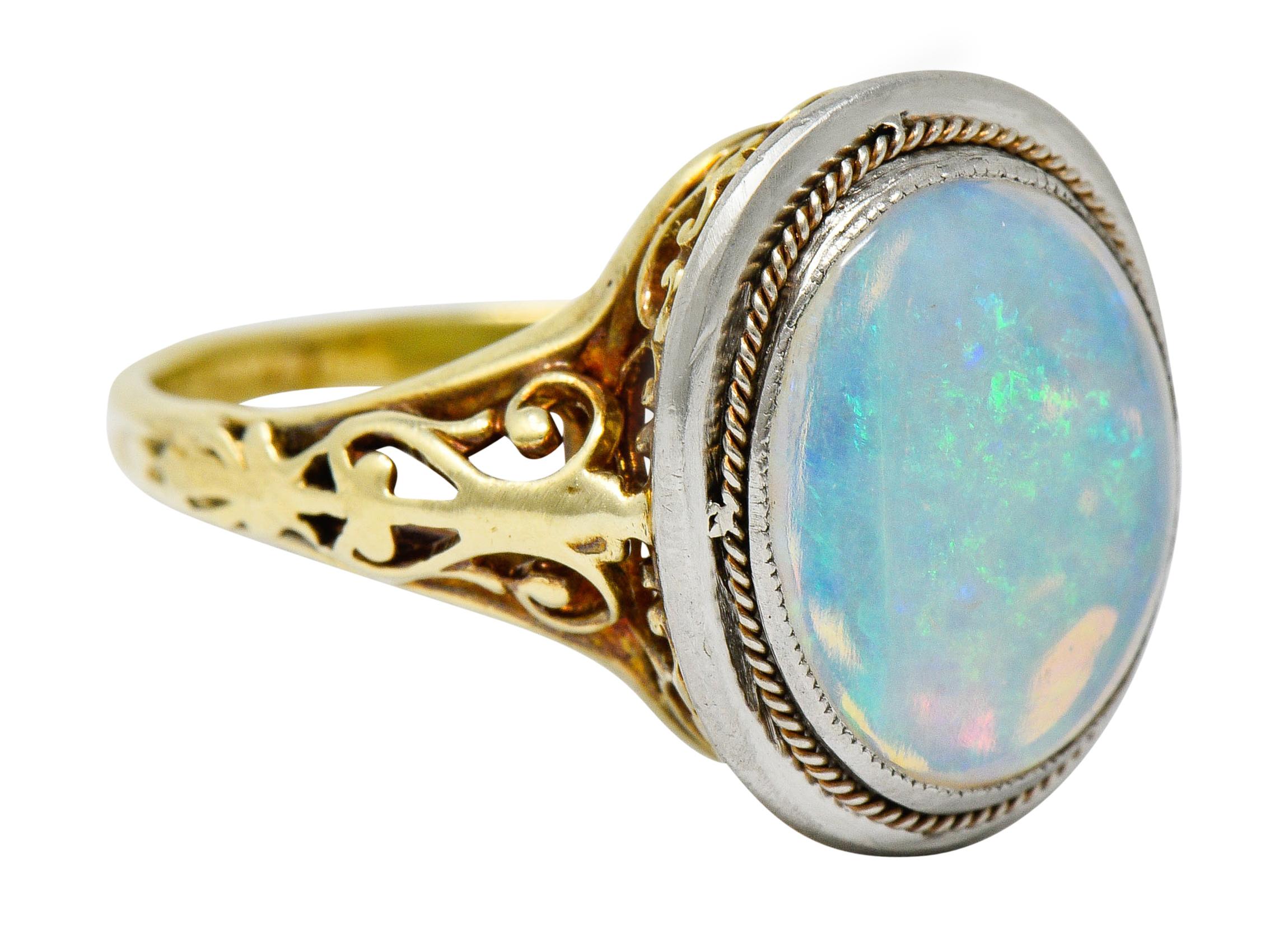 Centering an oval opal cabochon measuring approximately 11.5 x 10.0 mm

Translucent white color with strong green, yellow, blue play-of-color

Bezel set in a platinum surround accented by a gold twisted rope motif

Completed by scrolled filigree