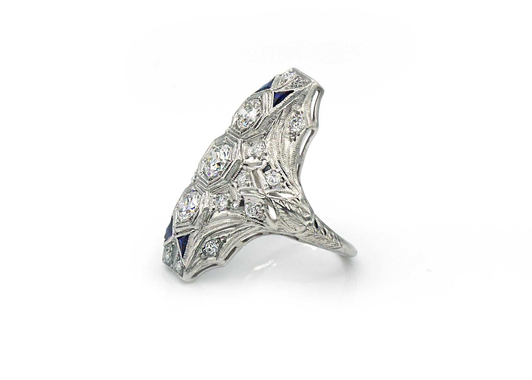 This early 20th Century antique platinum ring features 3 large old European cut diamonds at its center and 18 smaller diamonds accenting the openwork and filigree of the north-south central line. In addition, two bow-ties made of 4 deep blue