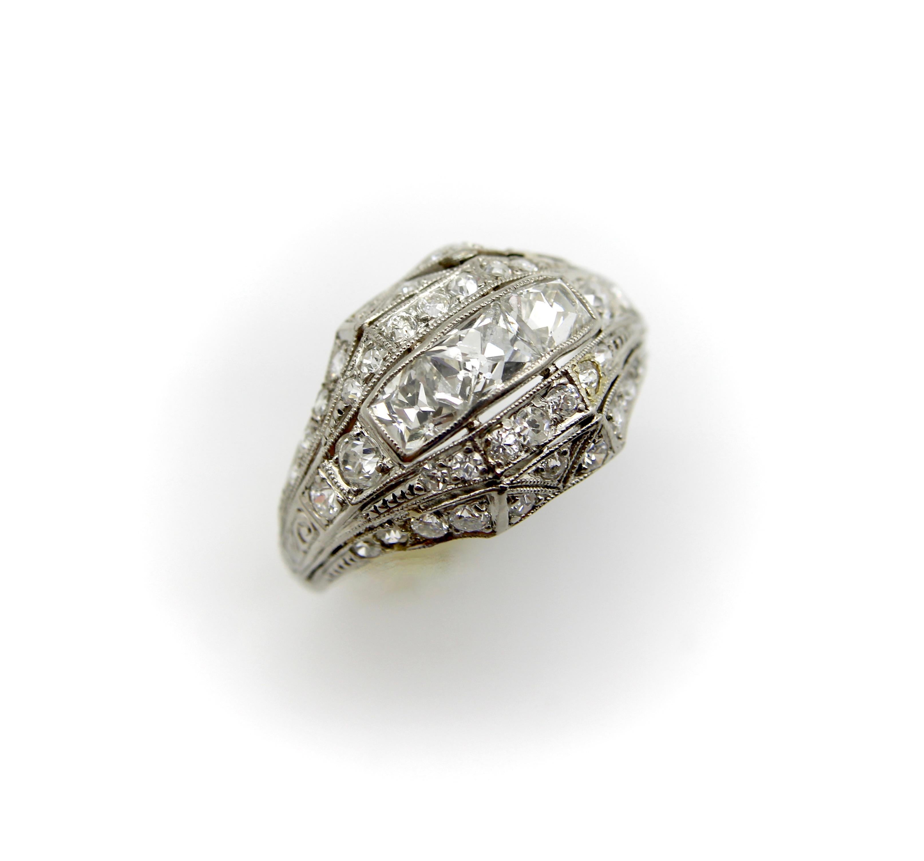Cette bague en platine sertie de diamants se situe à la charnière des époques édouardienne et Art déco. La pièce maîtresse de cette bague ornée est constituée de trois diamants Peruzzi taillés en chaton - l'un est de forme rectangulaire allongée,