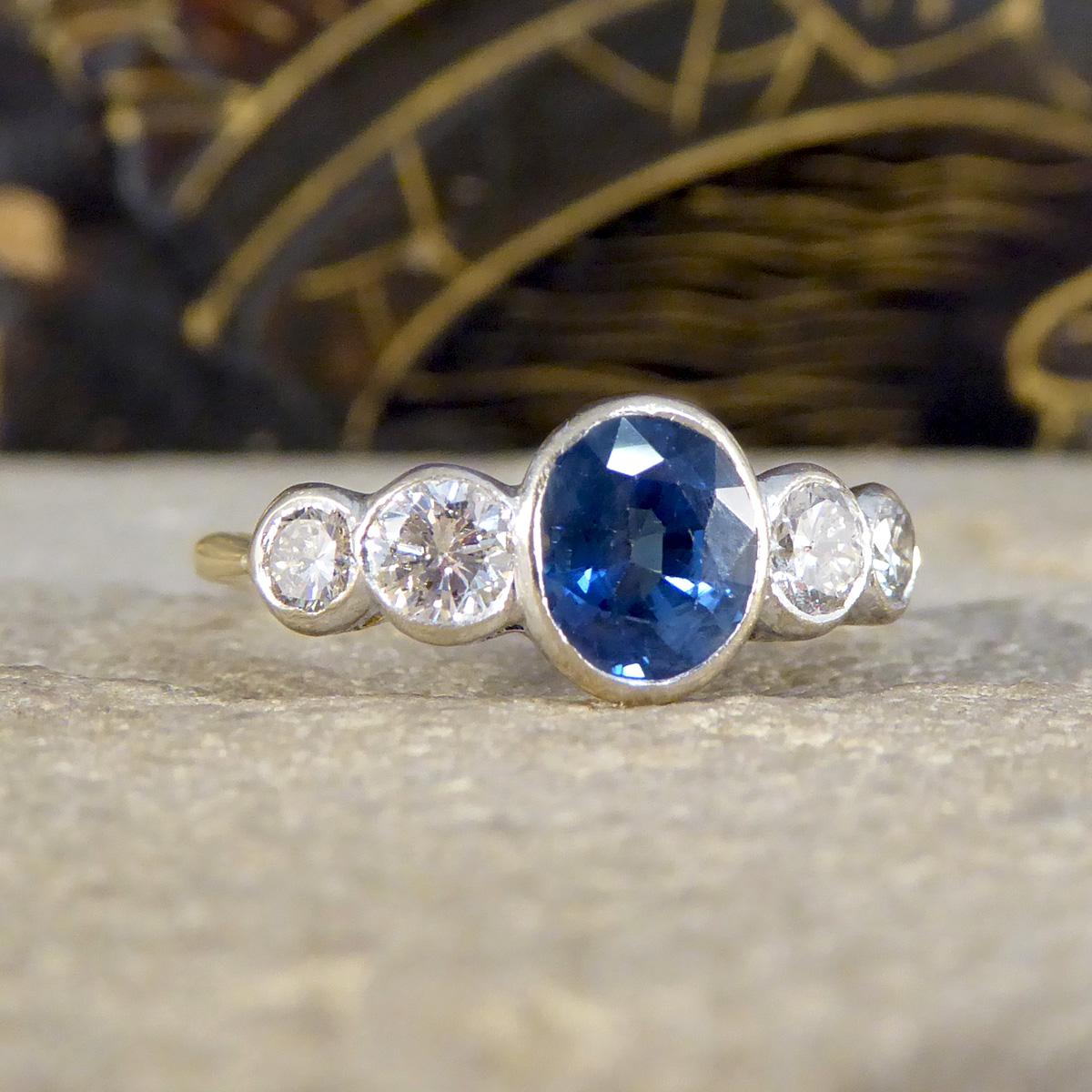 This Edwardian Sapphire and Diamond bezel set five stone ring is a testament to early 20th-century elegance. Crafted in 18ct white and yellow gold, it features a harmonious blend of one single deep blue sapphire in the centre weighing approximately