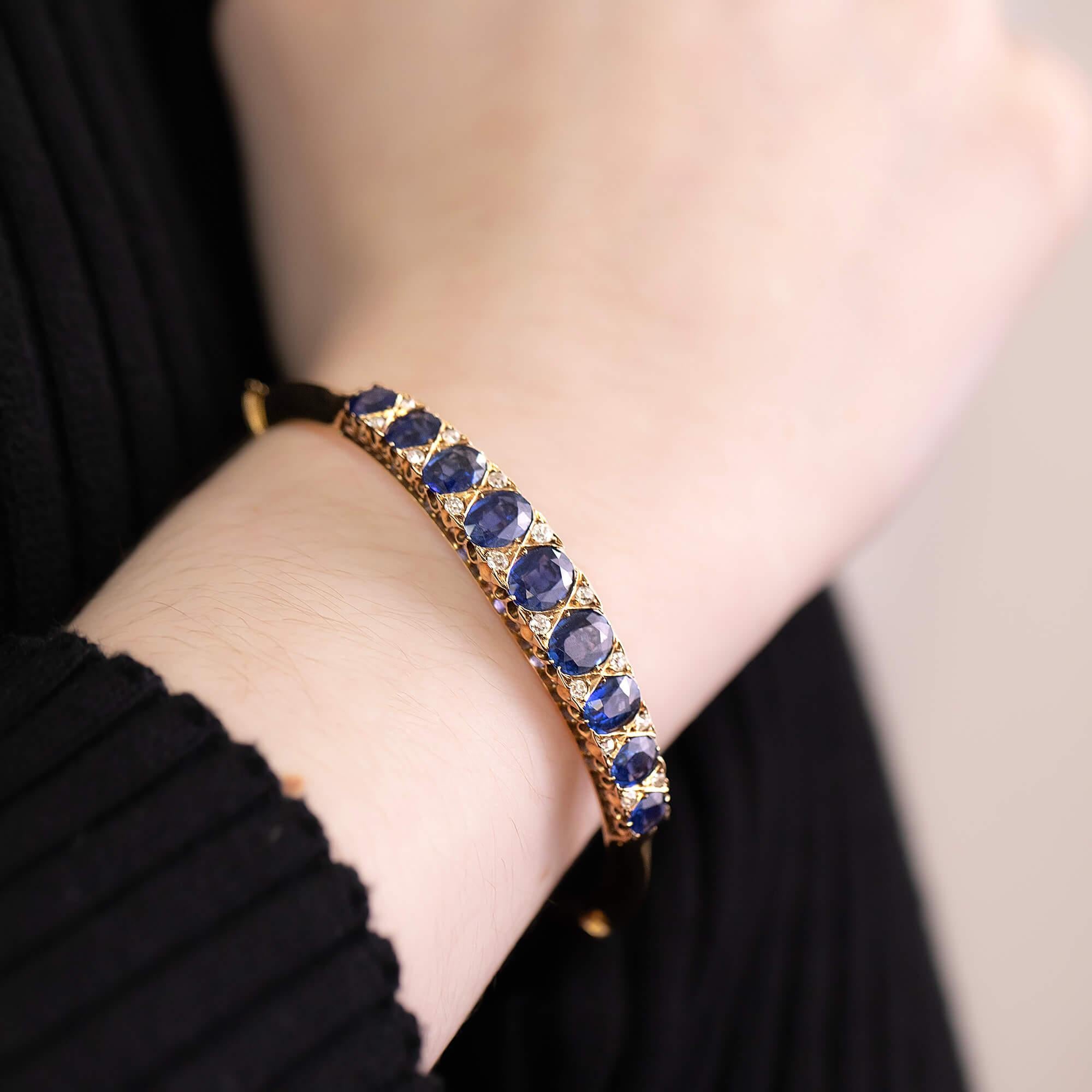 Edwardian sapphire and diamond-hinged bangle featuring beautiful gallery work and a safety chain.

Gemstone: Nine Oval-cut sapphires, grading from (4.85 x 3.9mm to 7.3 x 5.7mm), fine strong dark blue material, estimated total weight 7.50ct
Diamond: