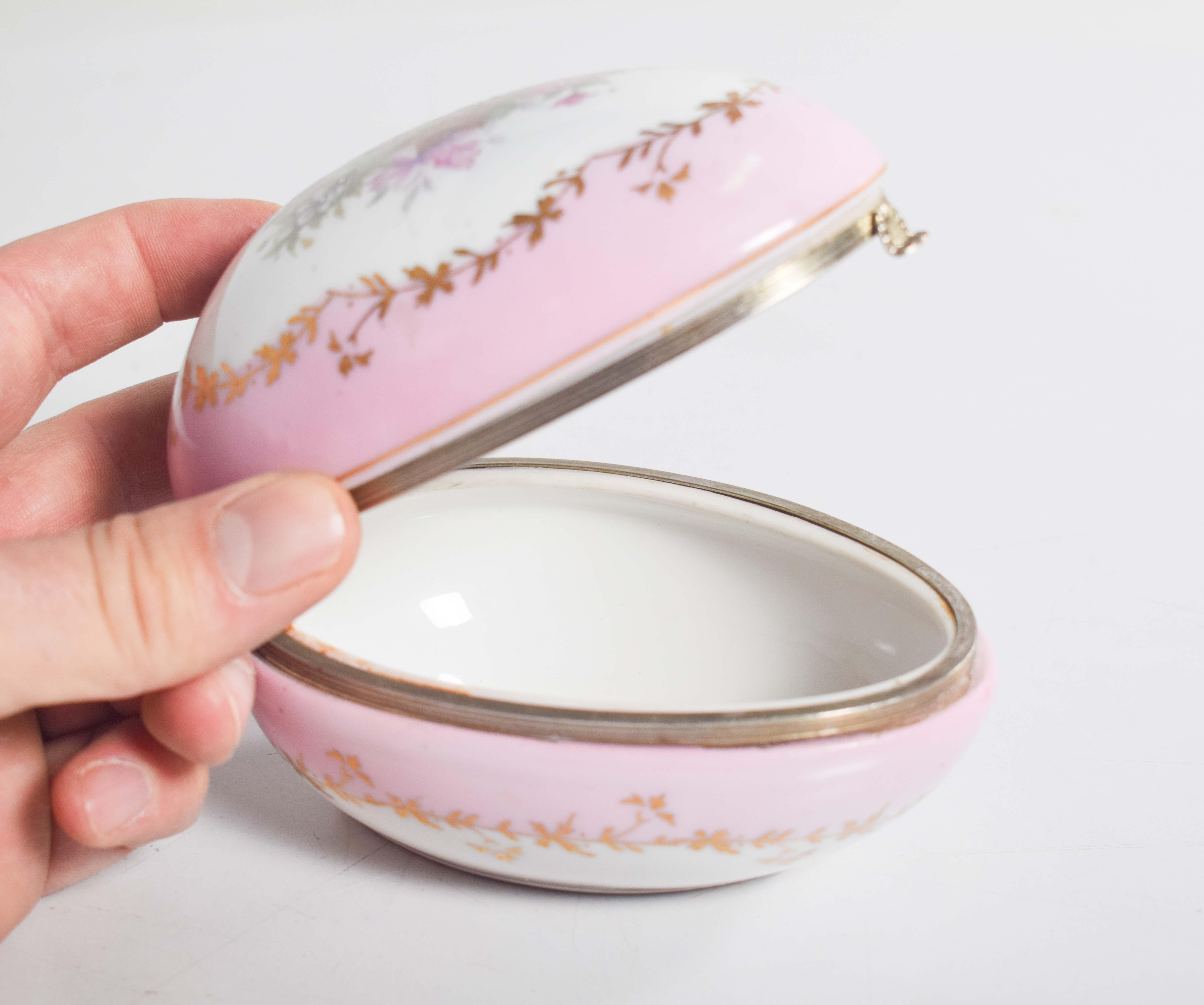 This delicate porcelain trinket box features an elegant, egg-shaped design, beautifully hand-painted and adorned with fine decorative details. The exterior showcases a soft pink band that wraps around the box, complemented by intricate floral motifs