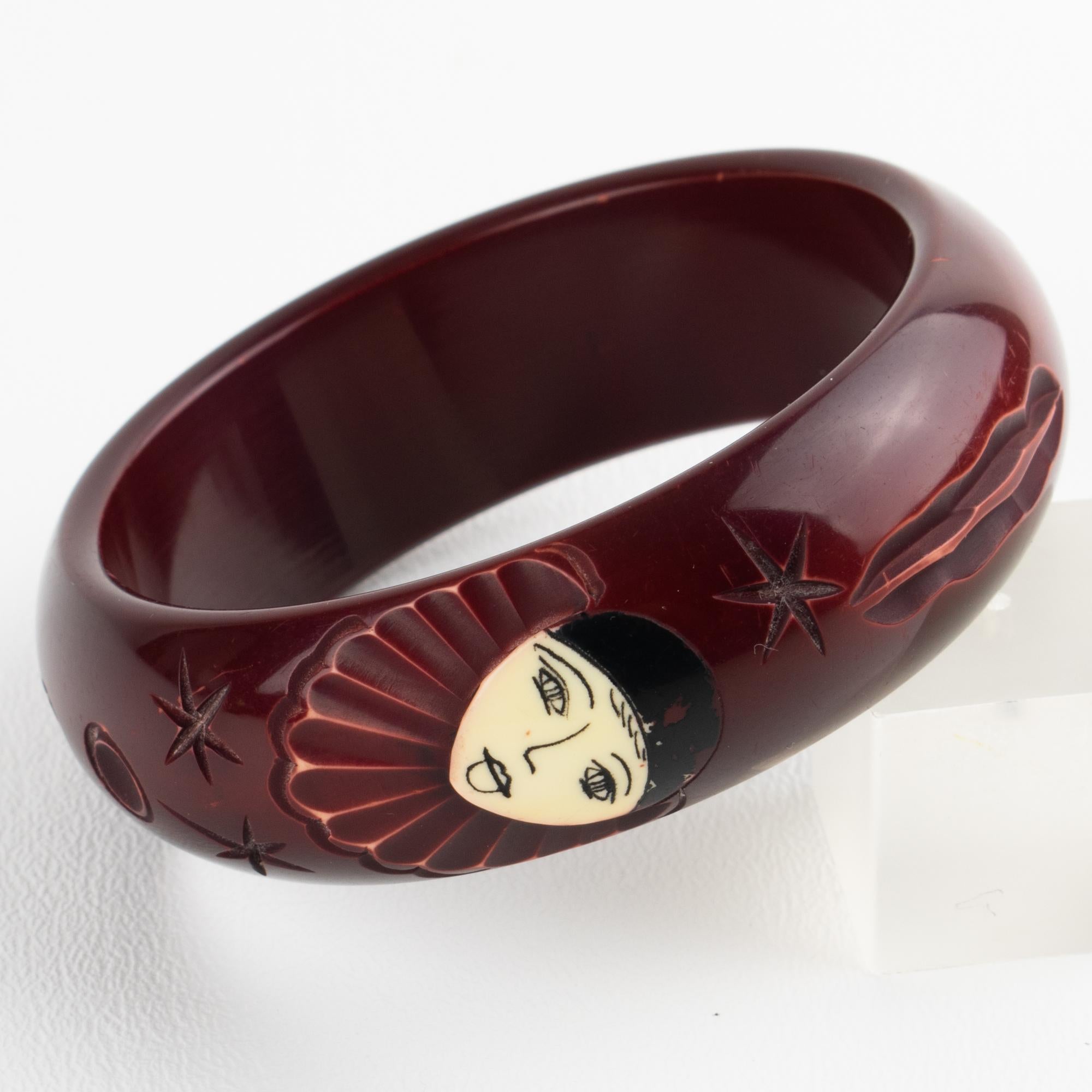 This is a rare Galalith bracelet bangle with a pierrot or clown carved design. The stunning piece features a chunky domed shape with deep eggplant purple color and a black and white contrasting carved design. The bracelet is adorned with a refined