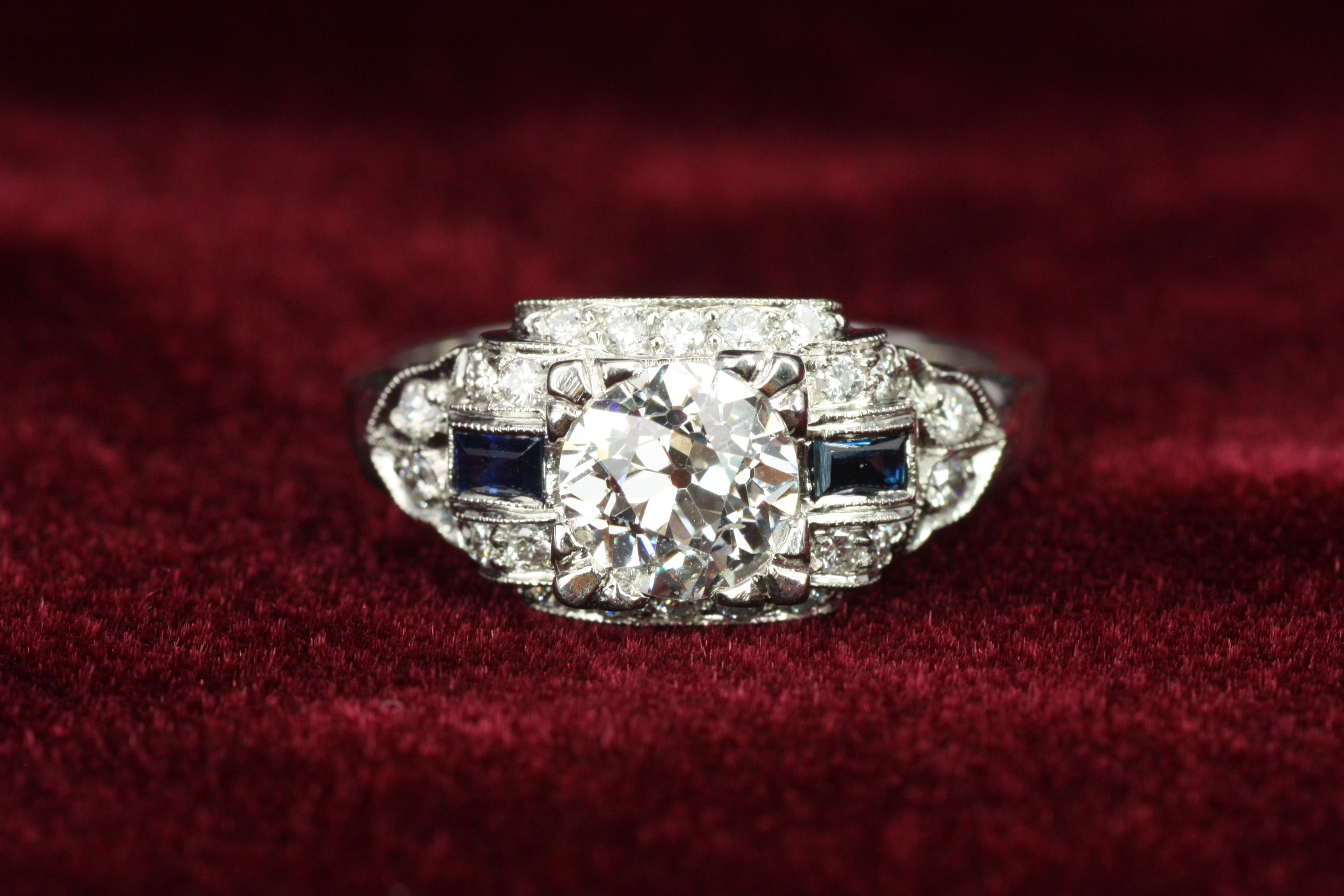 This Art Deco-inspired platinum ring captures vintage glamour with a modern touch. Featuring a 1.00ct old European cut center diamond and natural blue sapphires, its intricate design is both elegant and unique. A masterpiece for lovers of history