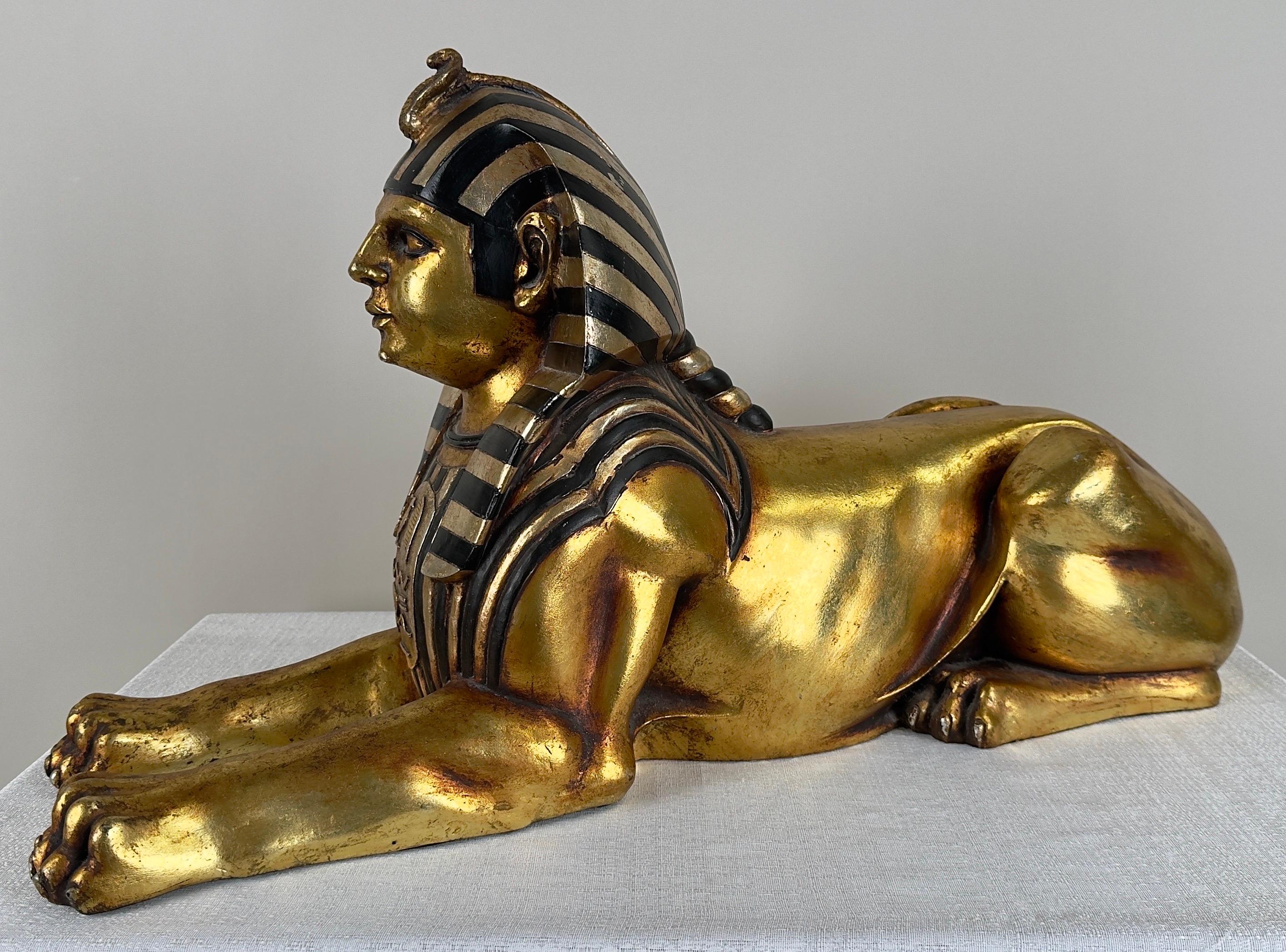 Una estatua de esfinge egipcia pintada a mano en oro radiante con detalles en negro. La esfinge, criatura mítica que simboliza la fuerza y la sabiduría, está representada en una pose clásica, con su alargado cuerpo felino y la regia cabeza de un