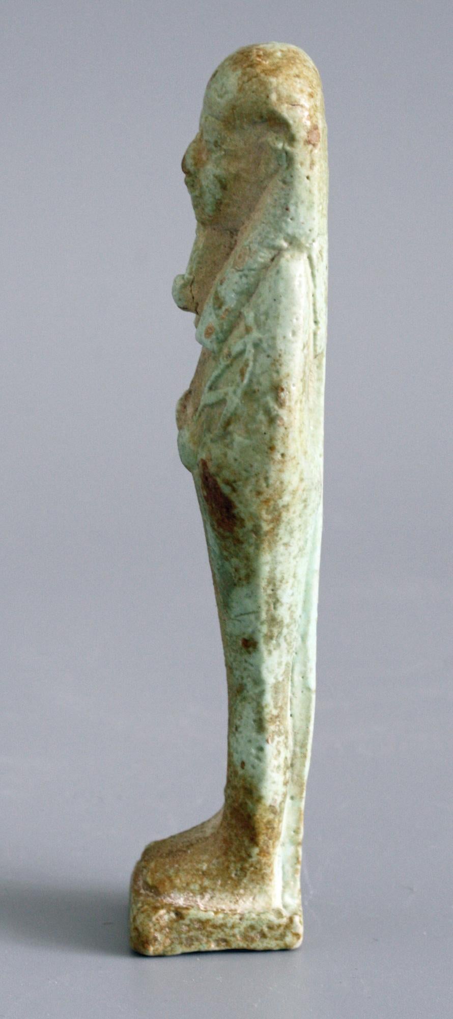 A very fine Egyptian pale green faience shabti figure believed to date from the Ptolemaic Dynasty 305-30BC. The figure is depicted in mummiform and wears a tripartite wig and false beard. He holds his arms on his chest holding a hoe and mattock with