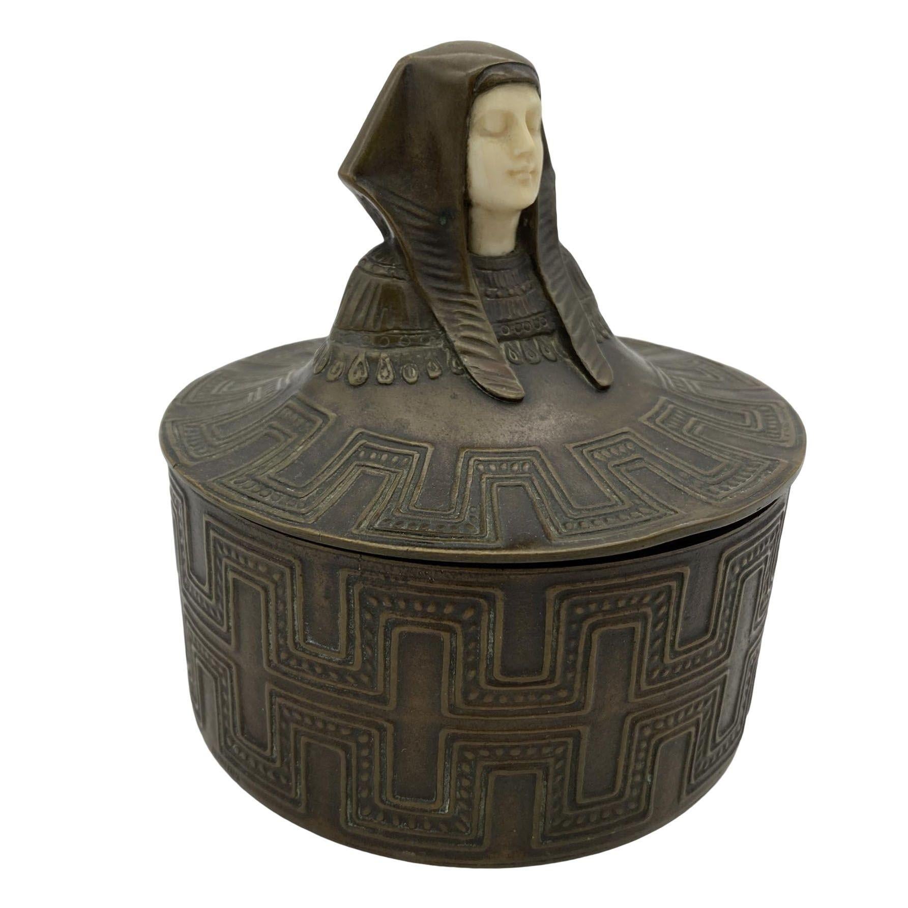 Plongez dans l'opulence de l'époque Art déco avec ce coffret en bronze coulé de style néo-égyptien. Orné d'une face en os méticuleusement sculptée, le couvercle amovible présente une figure royale de pharaon, qui se transforme en un relief