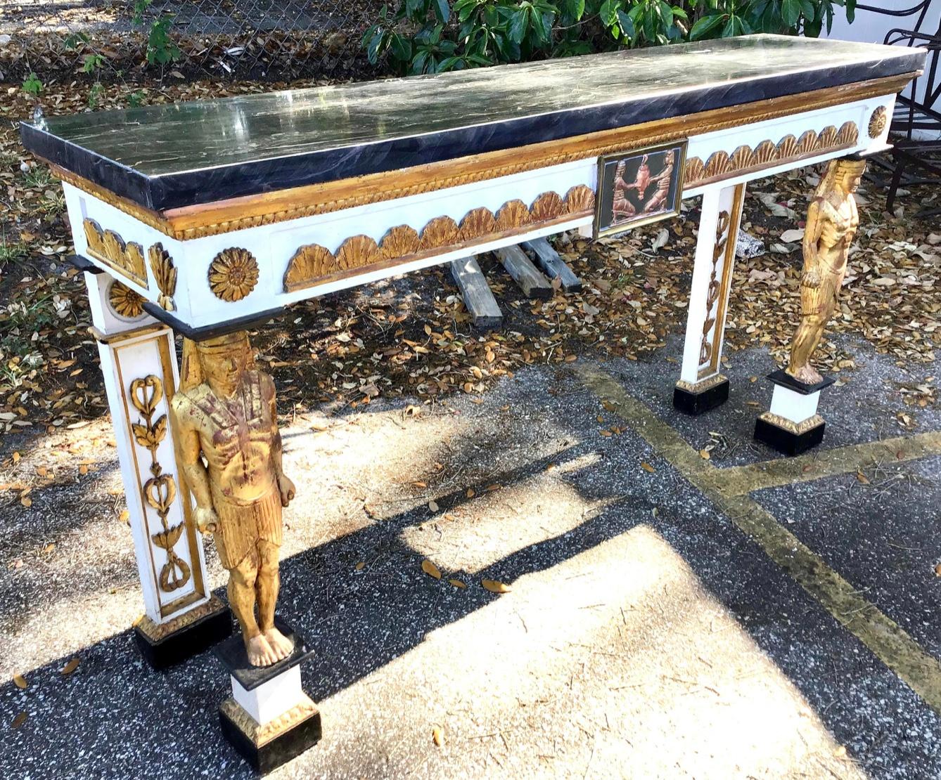 A large and impressive Egyptian Revival console table. Black portoro marble top. Frieze carved with gilt decorated fans and rosettes. Central medallion with gilt and framed Egyptian scene. The legs in the shape of standing gilt Sphinxes. A large and