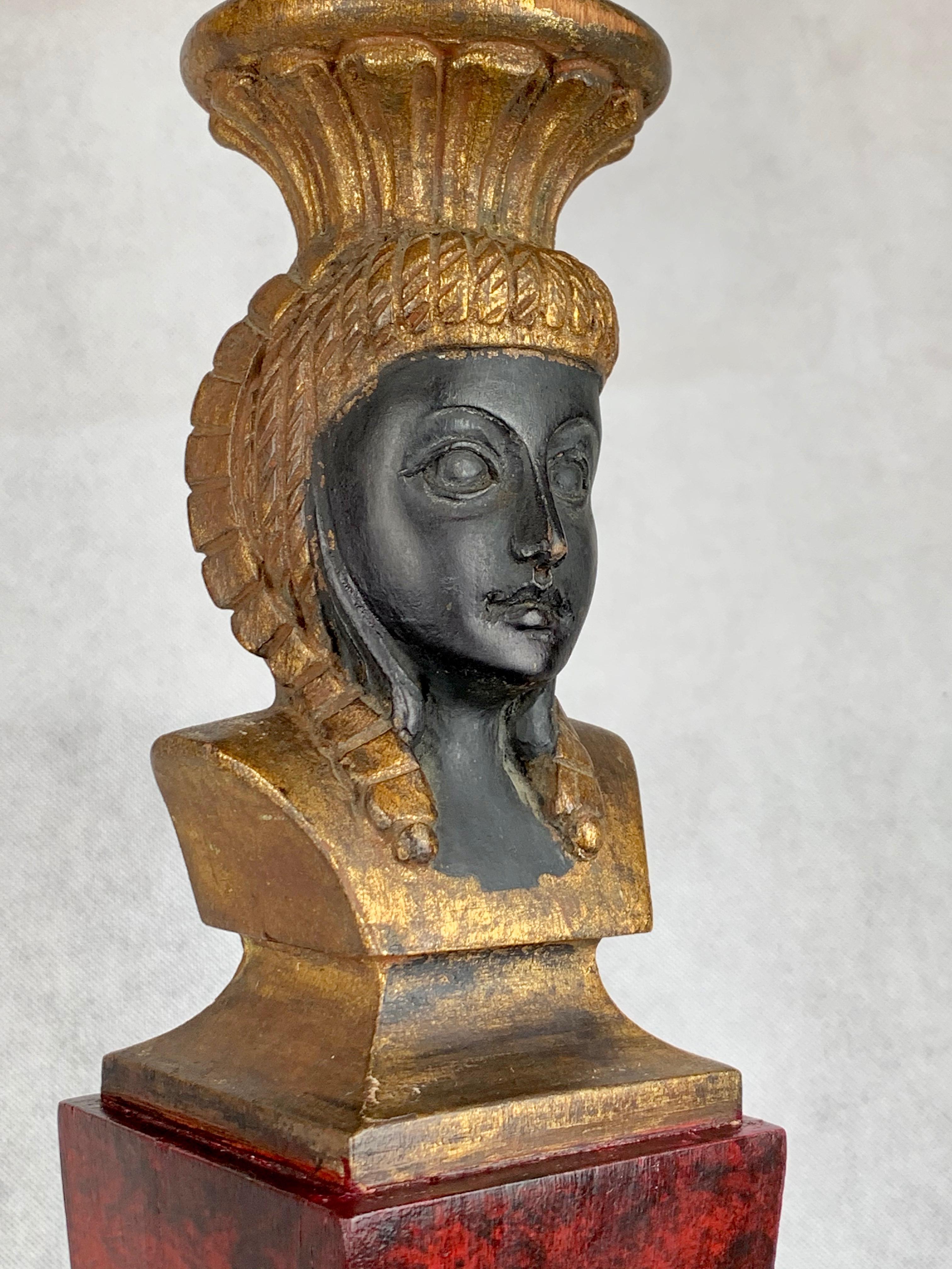 Lampe en bois sculpté de style égyptien. Le visage féminin ciselé et ébonisé est entouré d'une coiffe dorée. Le corps de la lampe est en faux marbre rouge profond, tandis que la base triple carrée est décorée de faux marbre rouge, de dorures et