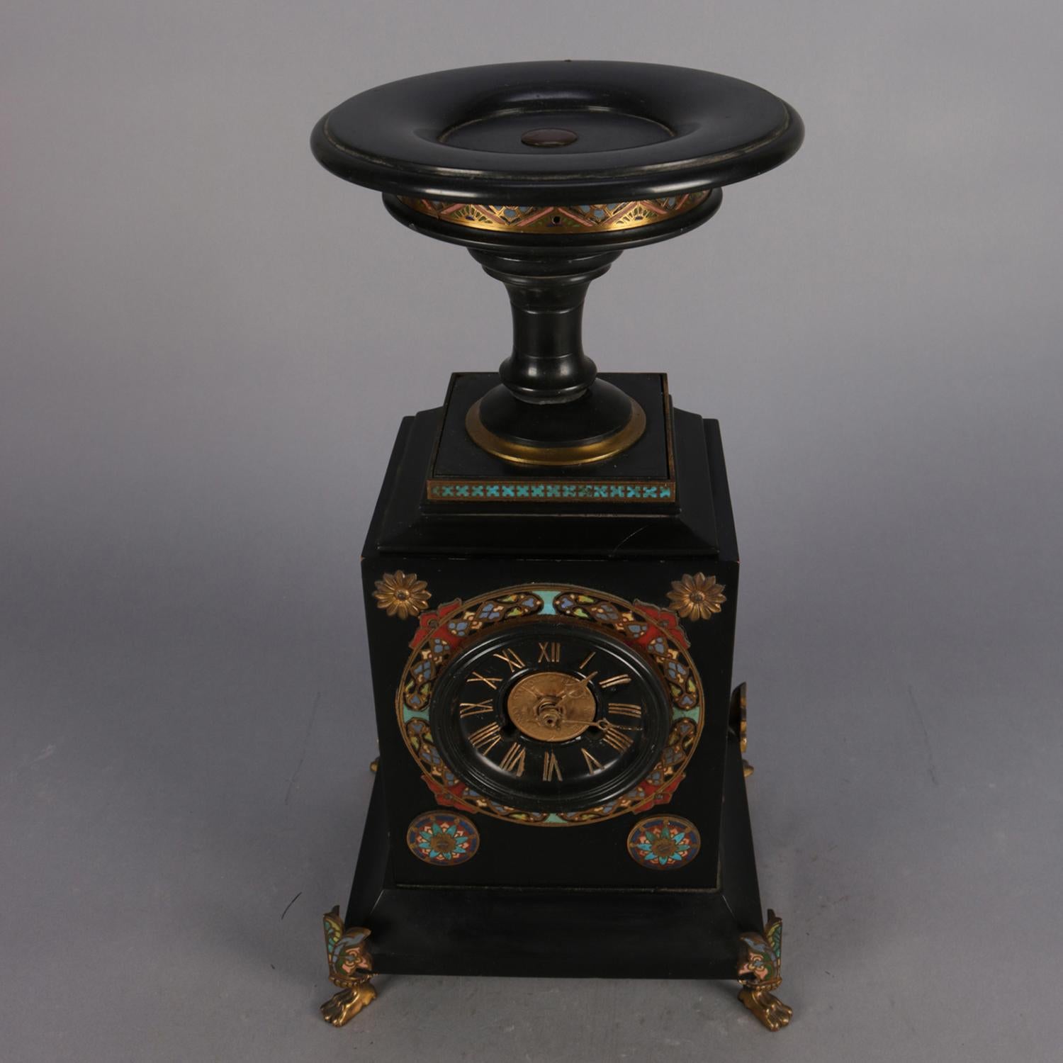 Egyptian Revival mantel clock features ebonized case with urn form compote finial, bronze attachments and enamelled decoration, face with Roman Numerals and central coin, sides having bronzed acanthus leaves and raised on bronzed paw feet, clock is