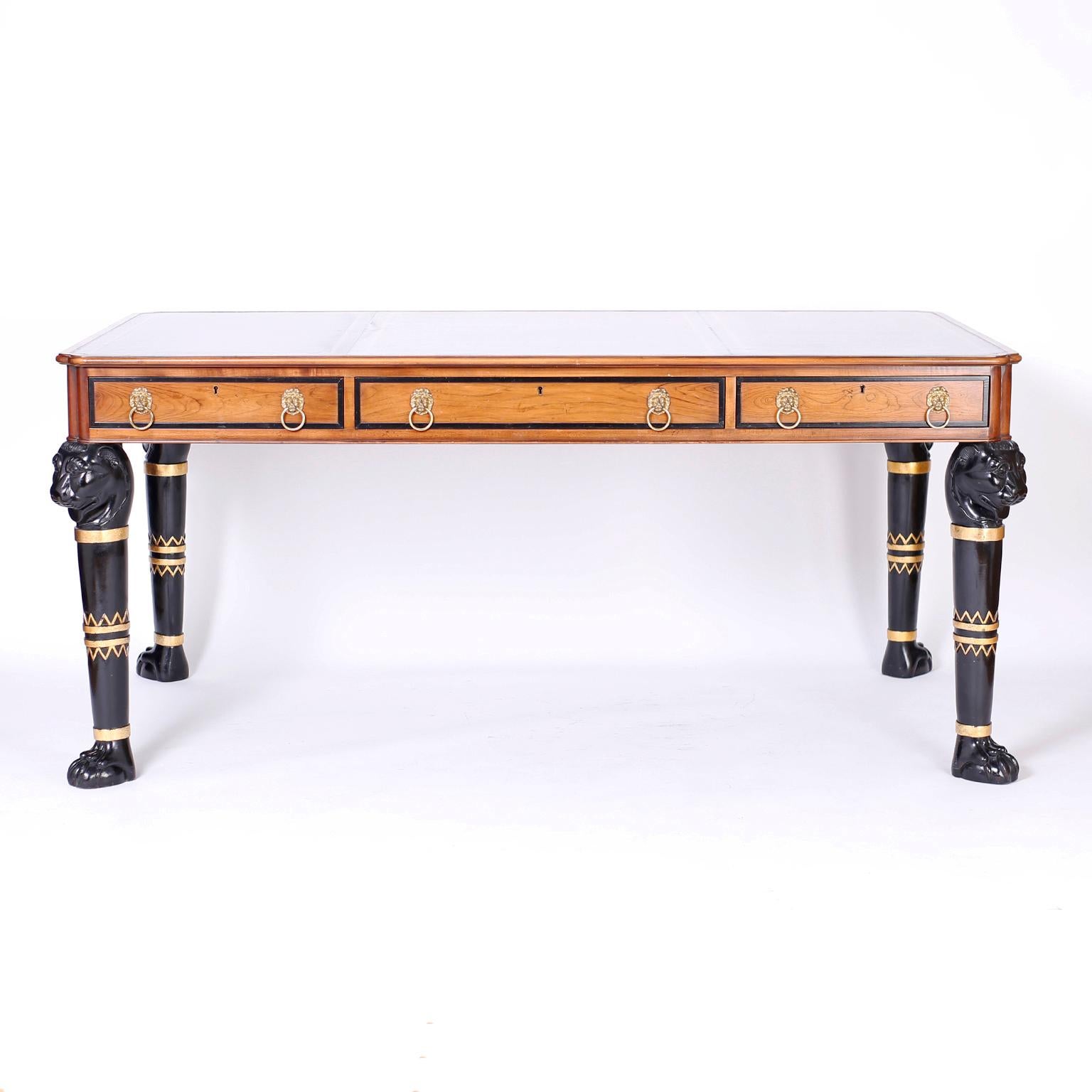 Egyptian revival or neoclassical desk or writing table crafted in walnut with three tooled leather panels on the top, three paneled drawers in front with brass lion head pulls and four ebonized legs decorated with distinctive Egyptian designs in