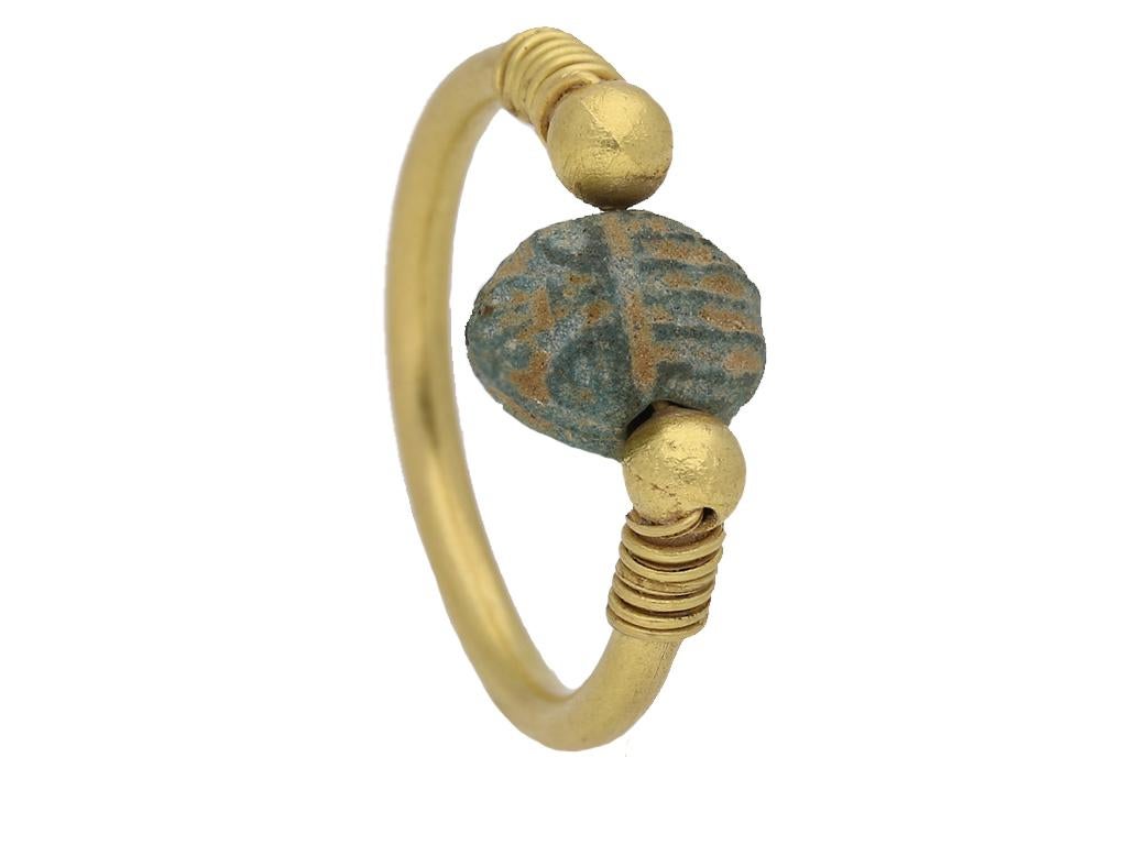 Egyptian scarab swivel ring. Set to centre with a finely modeled blue faience scarab bead, threaded through with a fine gold wire and positioned between two spherical terminals, proceeding to decorative wirework shoulders and culminating in a solid