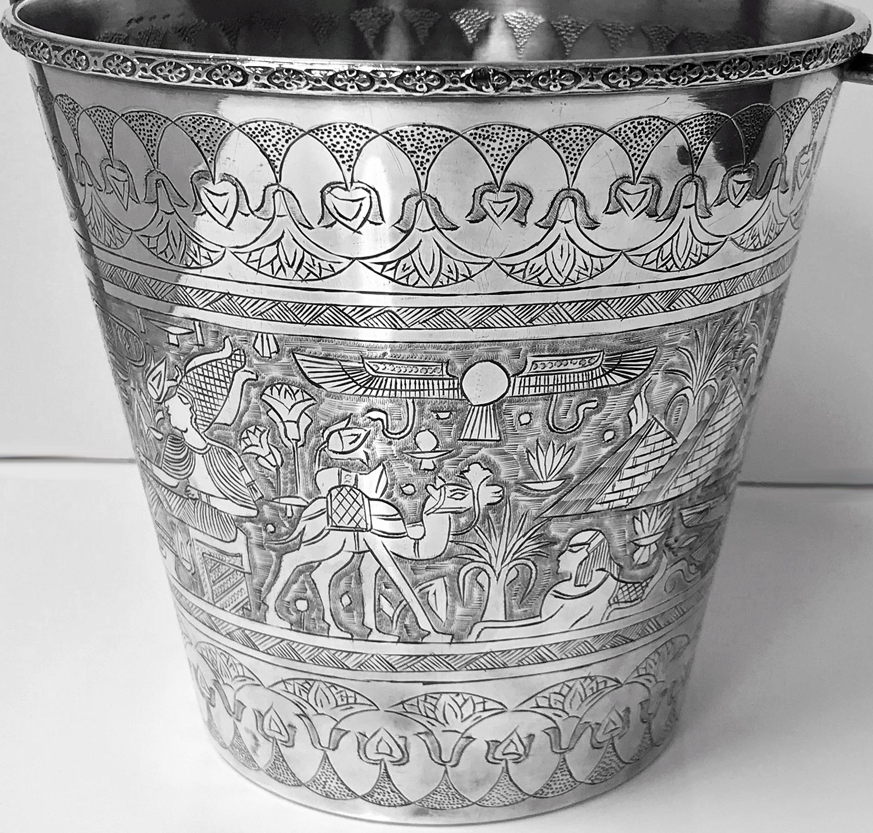 Egyptian silver wine bucket, circa 1920. The bucket of tapered form, richly engraved with multiple hieroglyphic depictions and pyramids, figures, horse’s chariots, snakes, serpents, camels, wing handle. Marks for Cairo under base. Measures: Height: