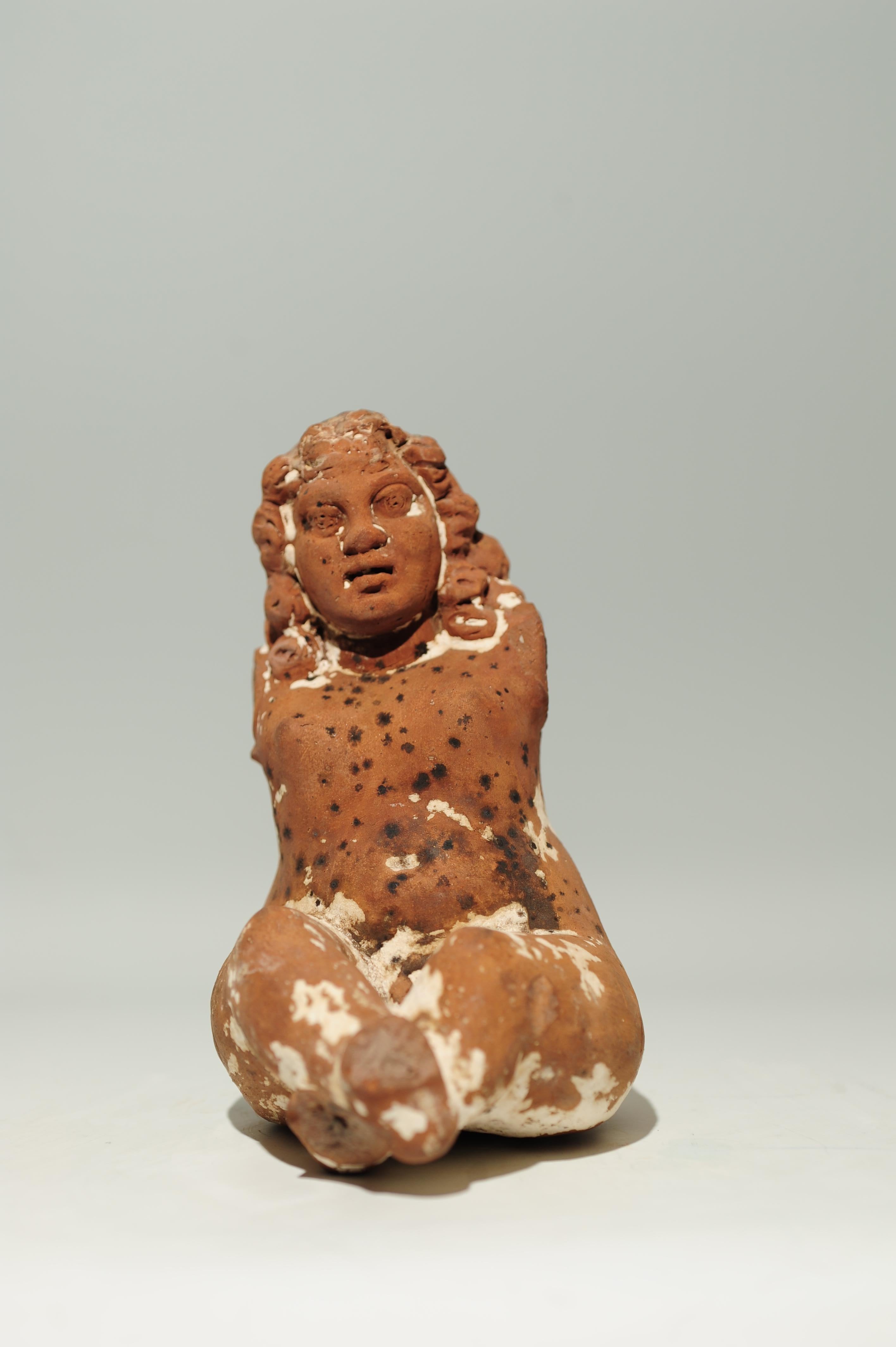 Molded. The stocky, bare boy sits with stretched, crossed legs on a surface that is no longer preserved today. The figure's head is turned slightly to the left. The shoulder-length wavy hair is reproduced as a compact mass. The back of the chubby