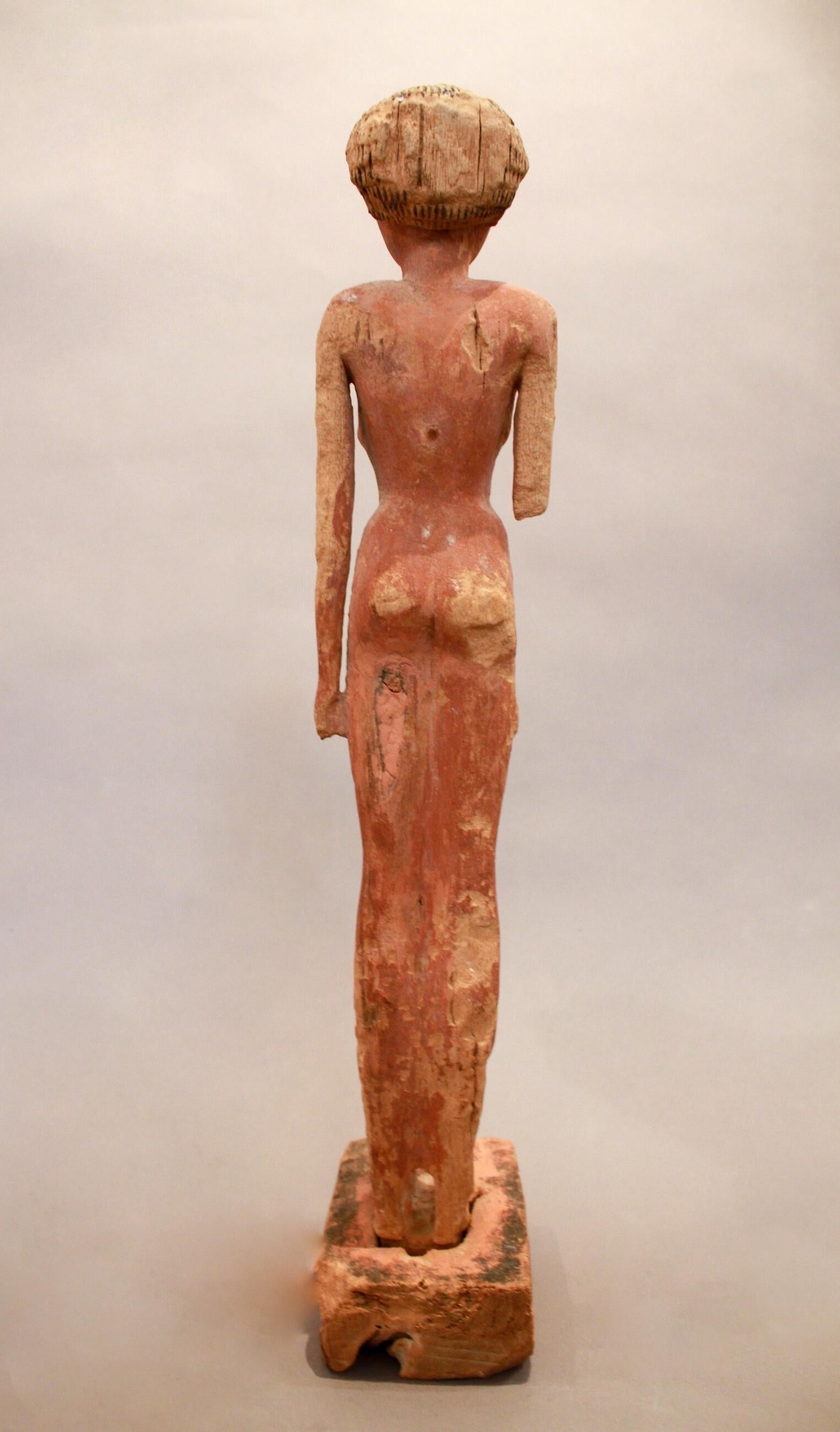 A statue of a nude women standing, with her arms laying alongside her body, wearing a wig that leaves the ears uncovered. The front of the wig cuts straight through the forehead, the eyes are painted white, surrounded by black lining to imitate